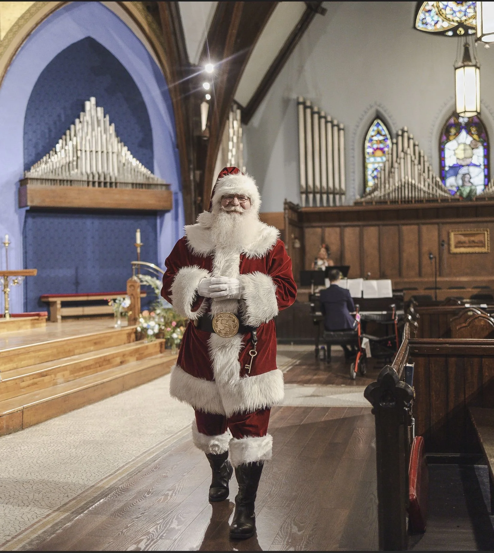 A person dressed as Santa Claus walking into a church with a wooden floor, stained glass windows, and pipe organ pipes in the background.