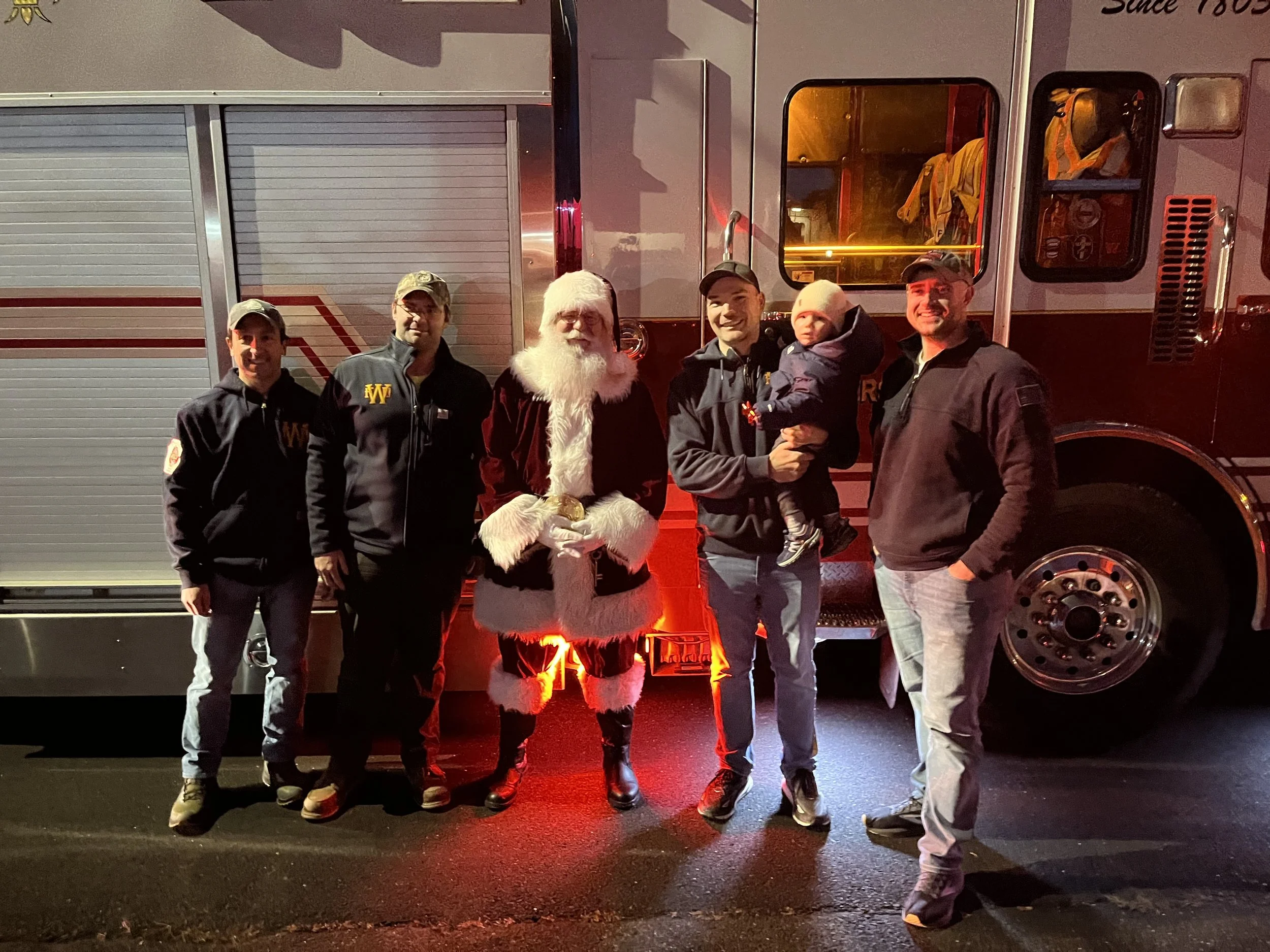 People standing in front of a fire truck, including a man dressed as Santa Claus and a man holding a small child, during nighttime with red lighting.