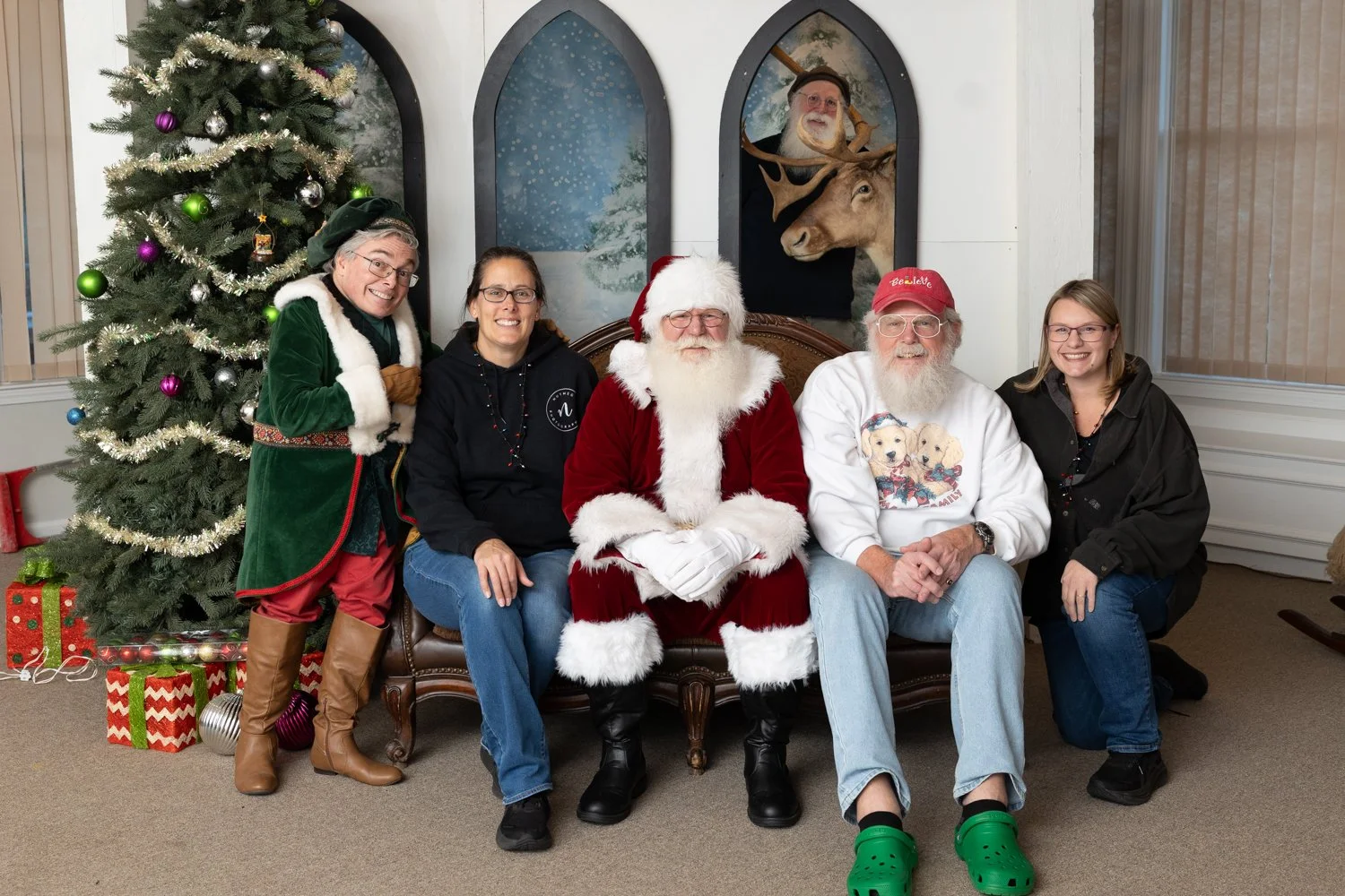 A group of six people, including Santa Claus, posing in front of a decorated Christmas tree with wrapped presents underneath. Two people are dressed in holiday-themed clothing, one with a Christmas sweater and red hat, and another in a green elf cost