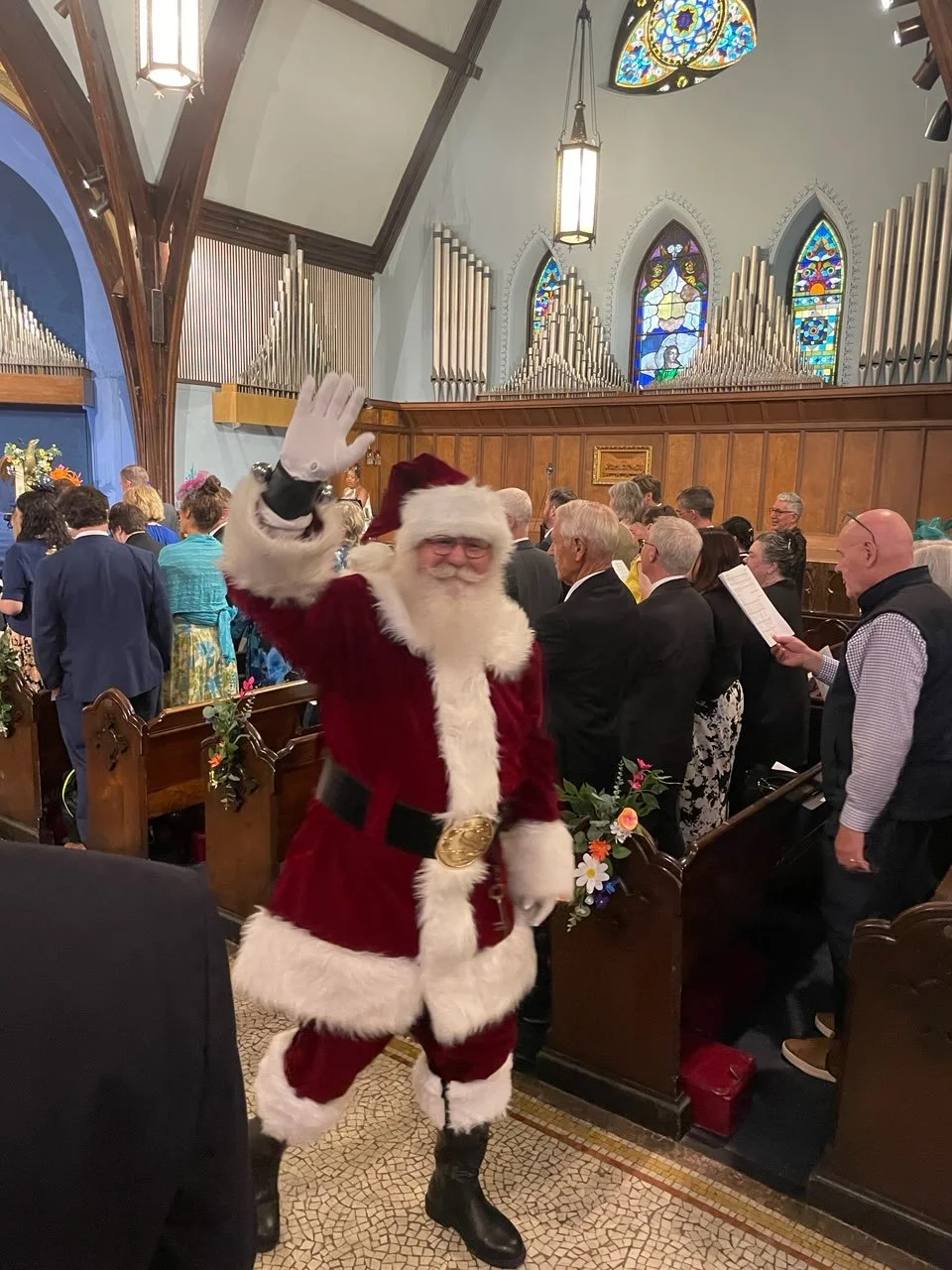 Santa Claus waving in a church during a Christmas event, with people gathered in pews and stained glass windows in the background.