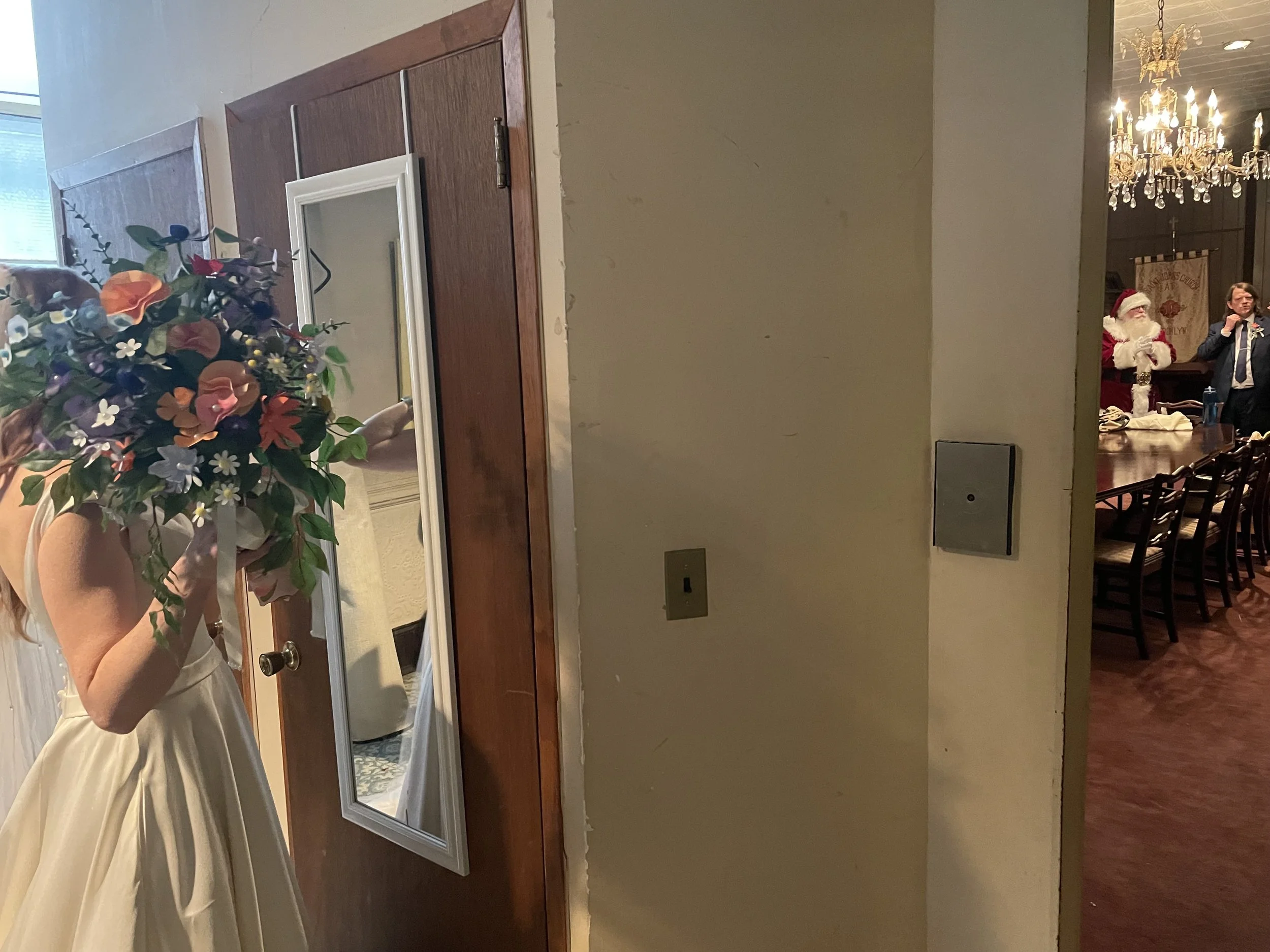 A woman in a white dress taking a mirror selfie in a room, holding a bouquet of colorful flowers. In the background, a chandelier and a man dressed as Santa Claus are visible, along with a woman talking on the phone.