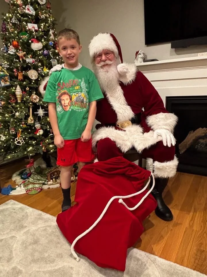 A young boy in a green shirt and red shorts standing next to Santa Claus, who is sitting by a decorated Christmas tree, with a large red sack of presents in front.