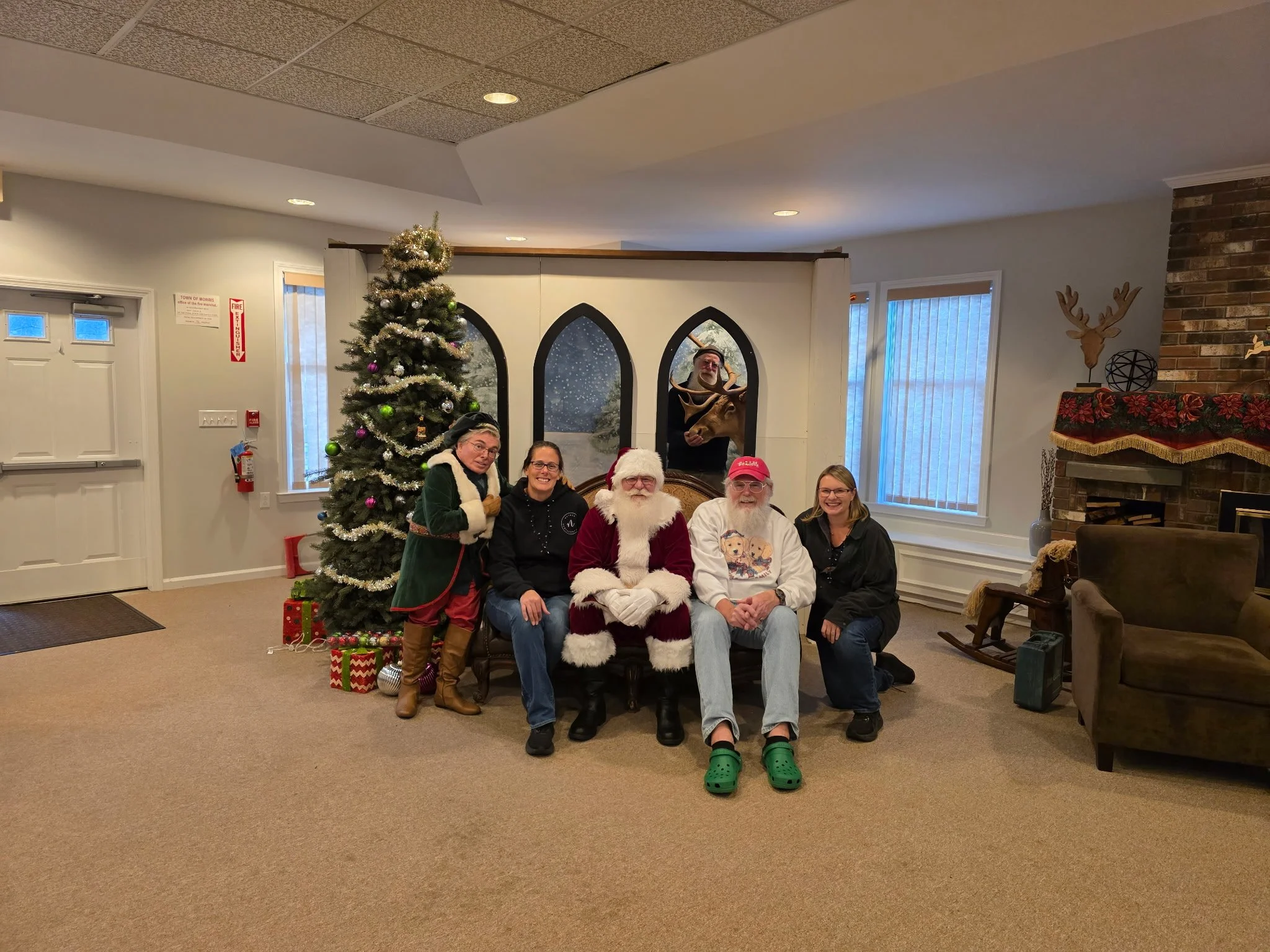 People celebrating Christmas with Santa Claus, sitting in front of a decorated Christmas tree with gifts, in a festive room.