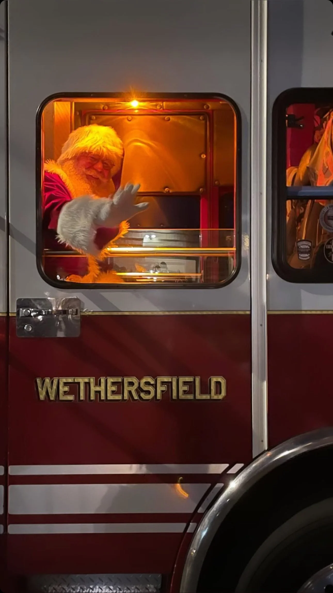 A person dressed as Santa Claus waving from a fire truck with roofing letters spelling 'Wethersfield' on the side.
