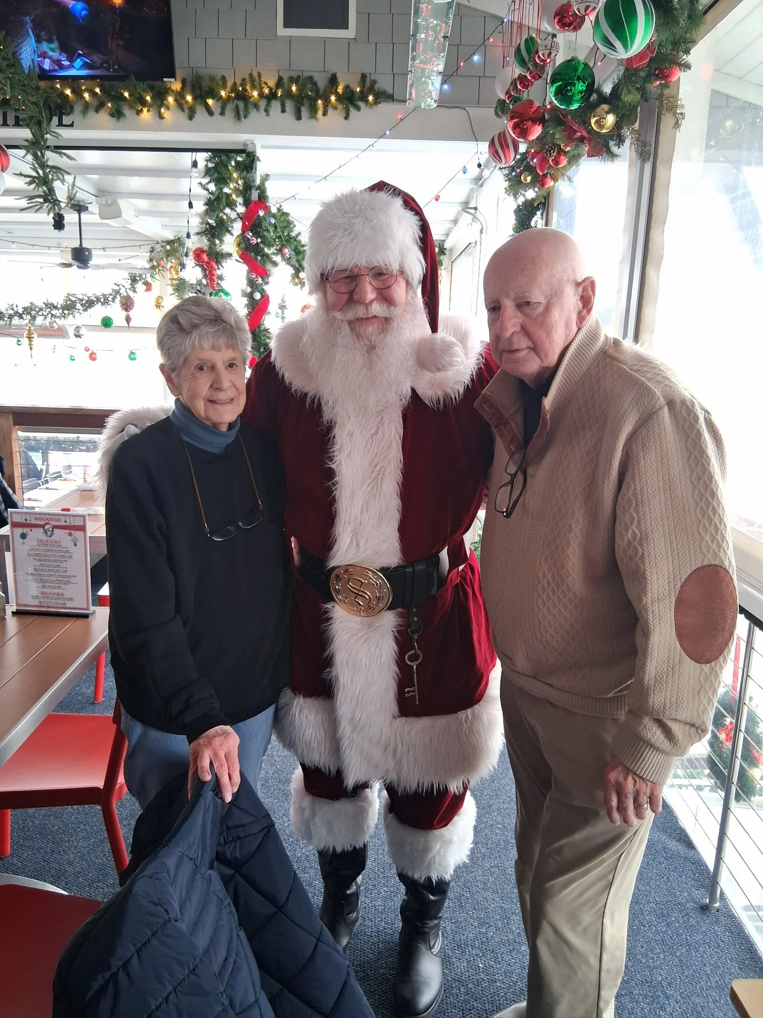 Three elderly individuals with festive holiday decorations, including a person dressed as Santa Claus, inside a decorated space with Christmas ornaments and lights.