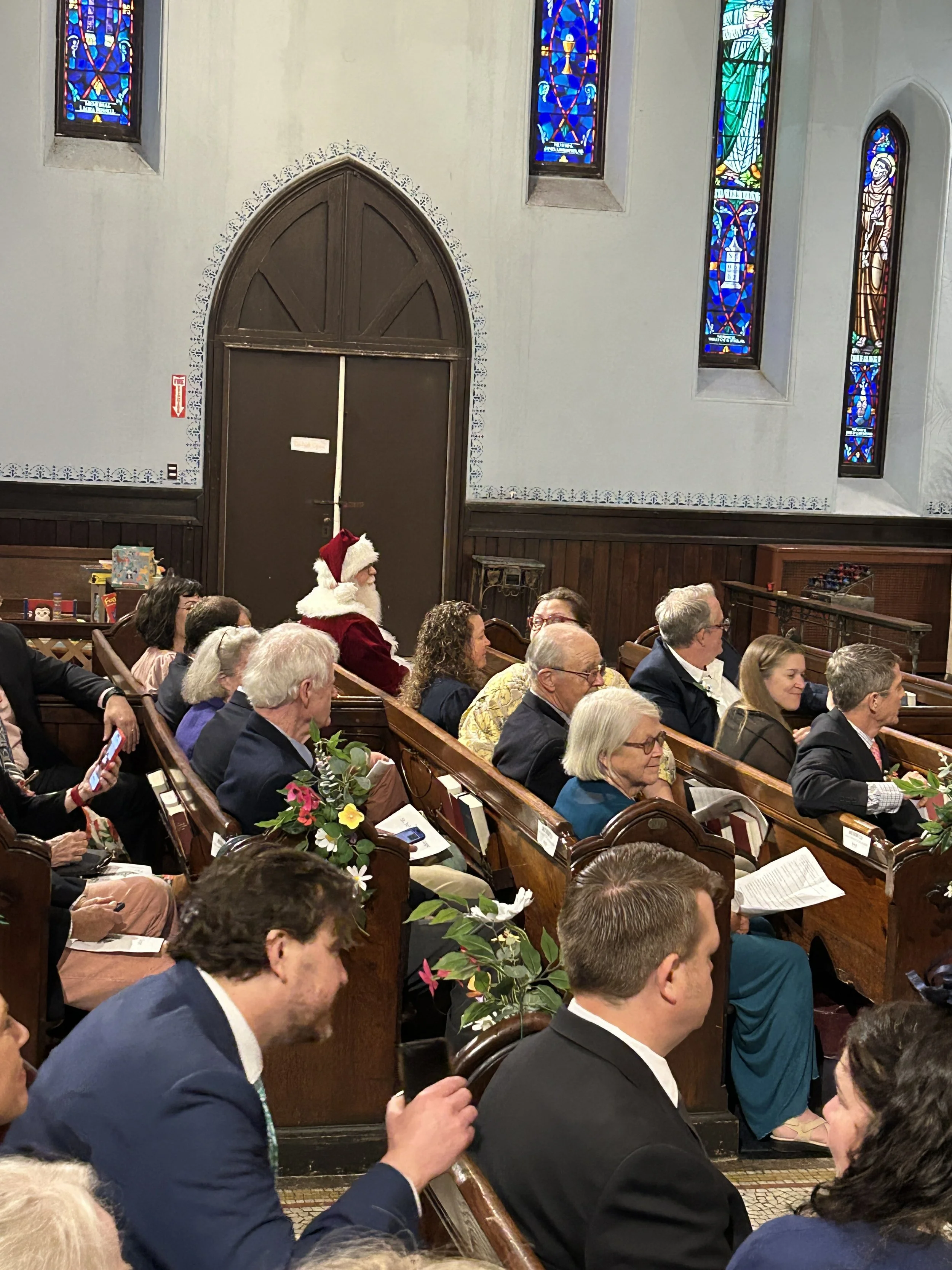 A group of people attending a church service, with some wearing formal attire, sitting on wooden pews. One person dressed as Santa Claus is present among the attendees. The church has white walls, stained glass windows, and wooden accents.