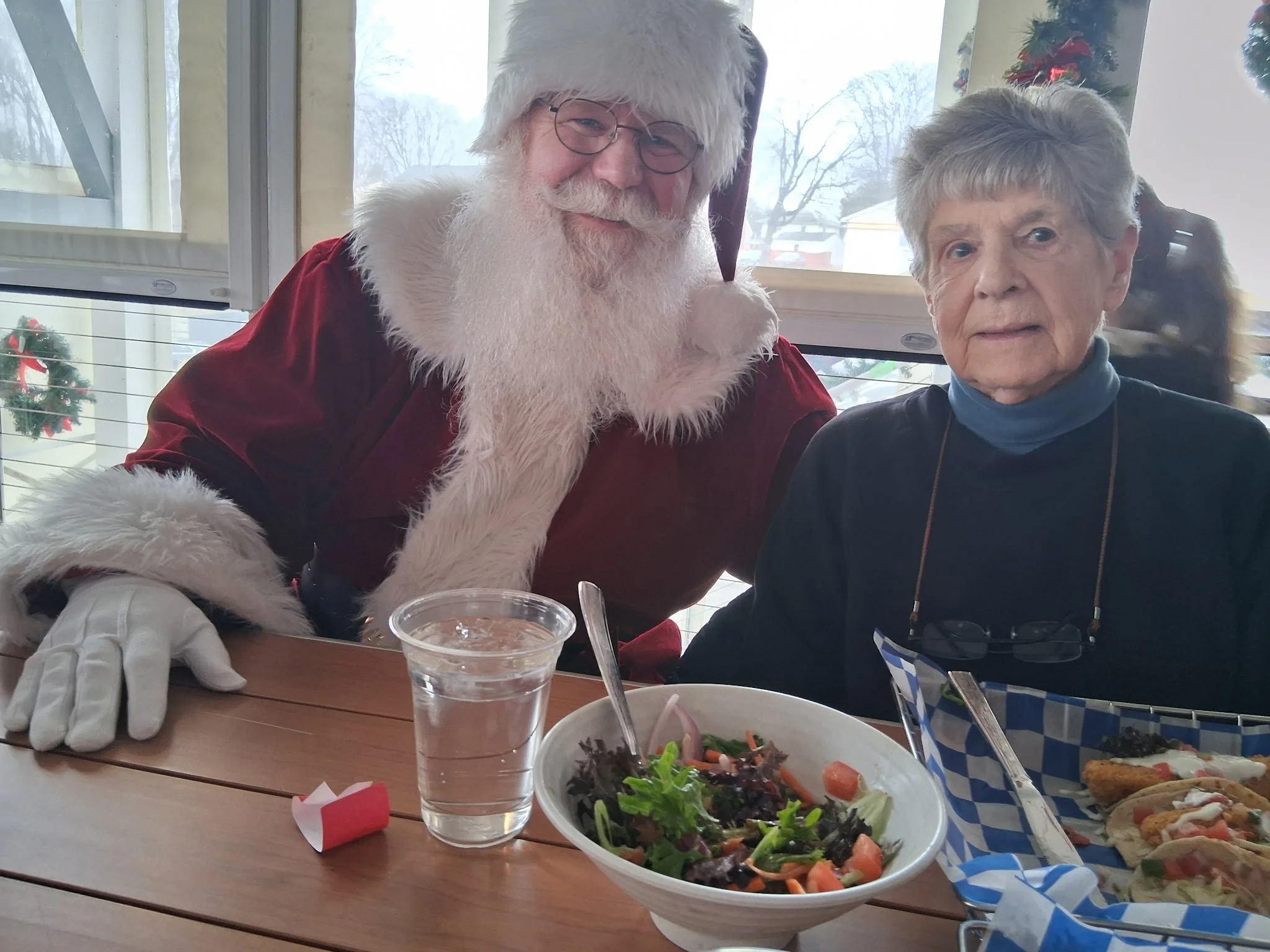 A man dressed as Santa Claus and a woman are sitting together at a table with food and drinks, with Christmas decorations in the background.