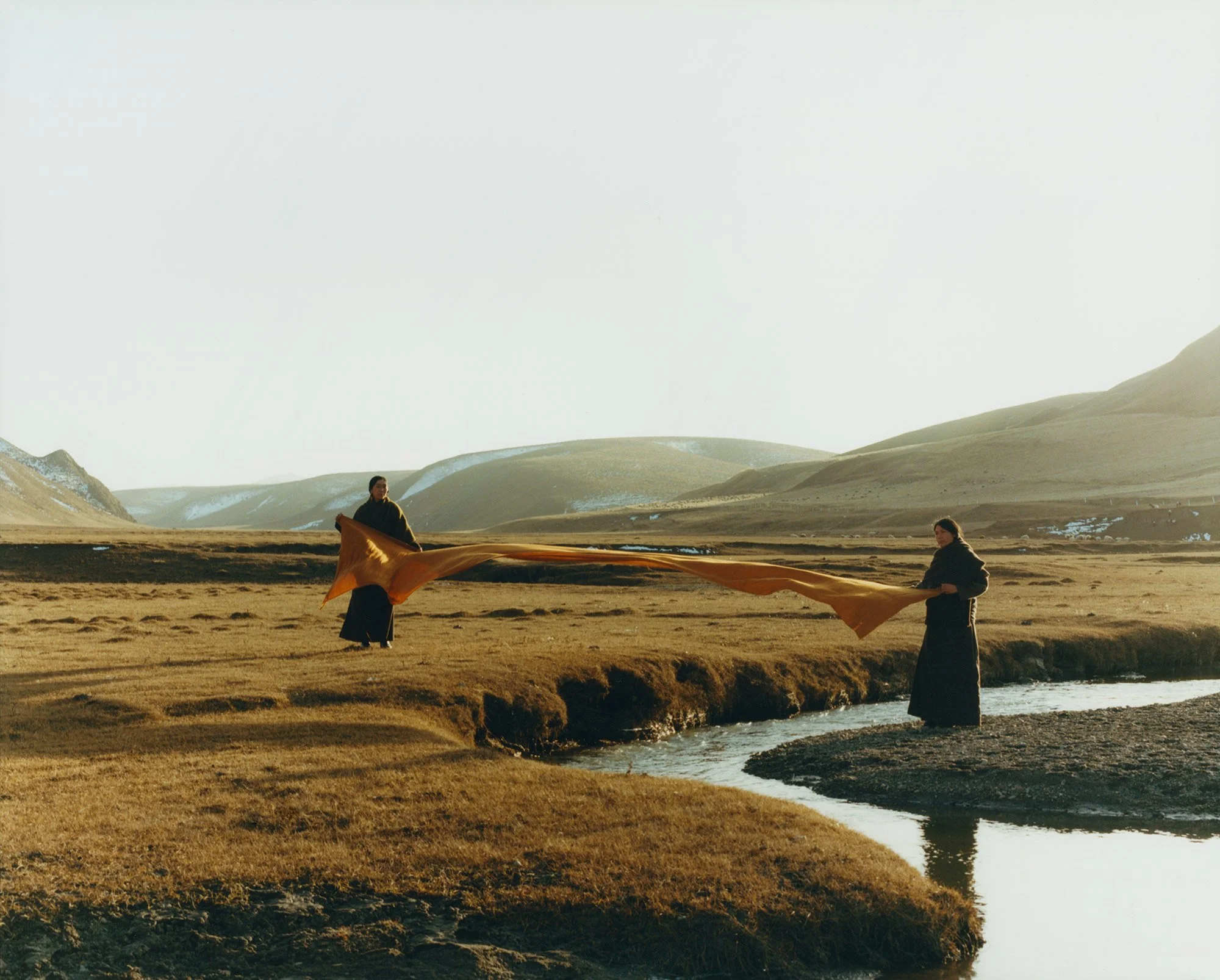 Two people. standing on opposing sides of a river, ruffling out a piece of long cloth.