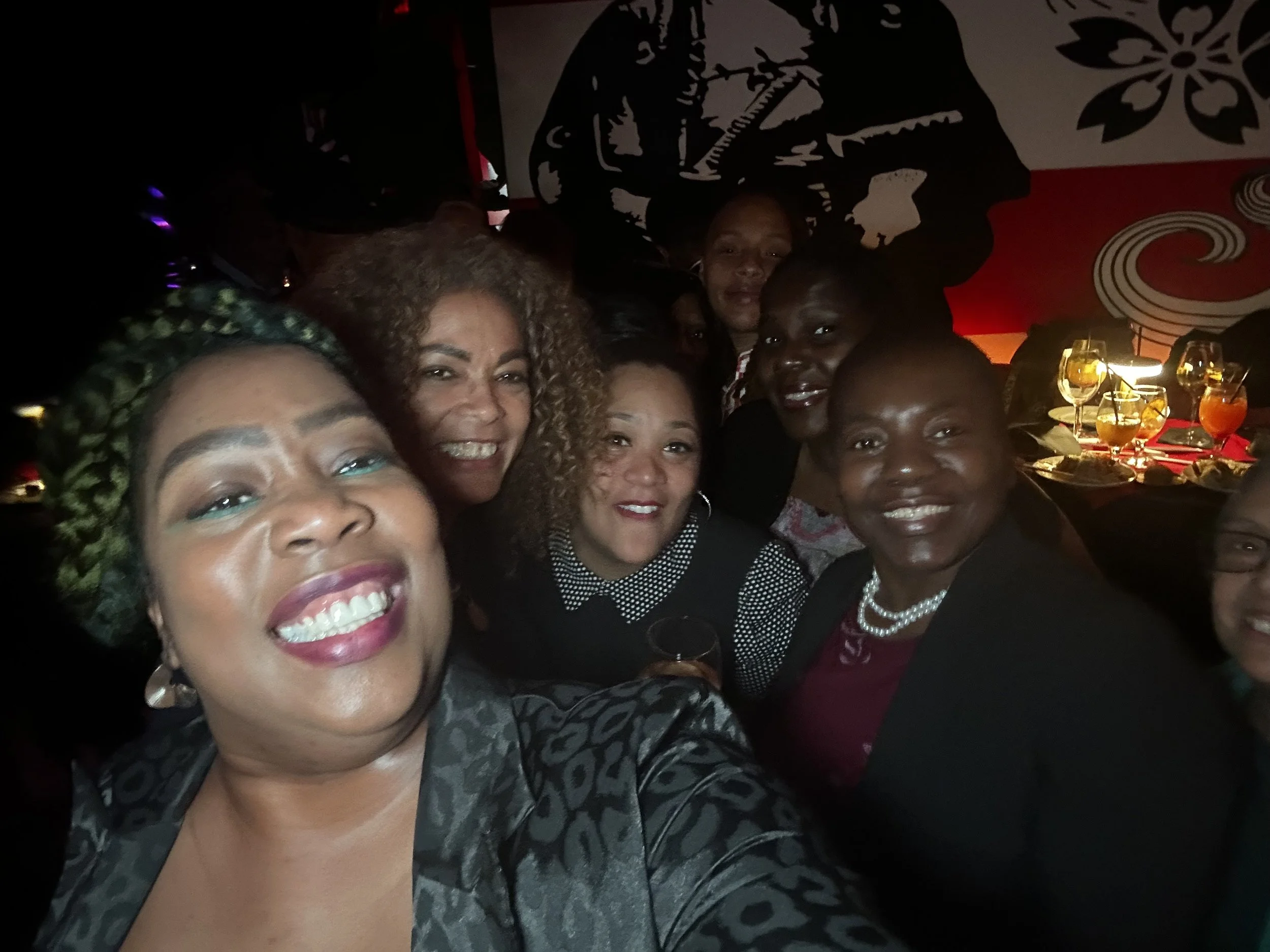 Group of women smiling and taking a selfie at a celebration or gathering in a dimly lit setting with decorated tables and drinks in the background.