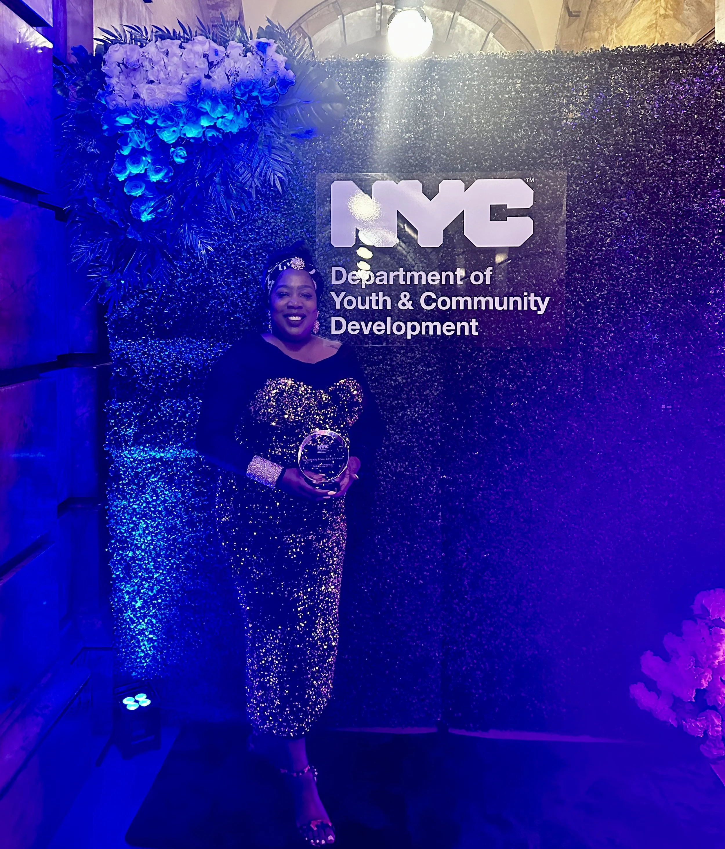 A woman standing in front of a decorated backdrop for the NYC Department of Youth & Community Development, wearing a black sequined outfit and holding a clear circular award.