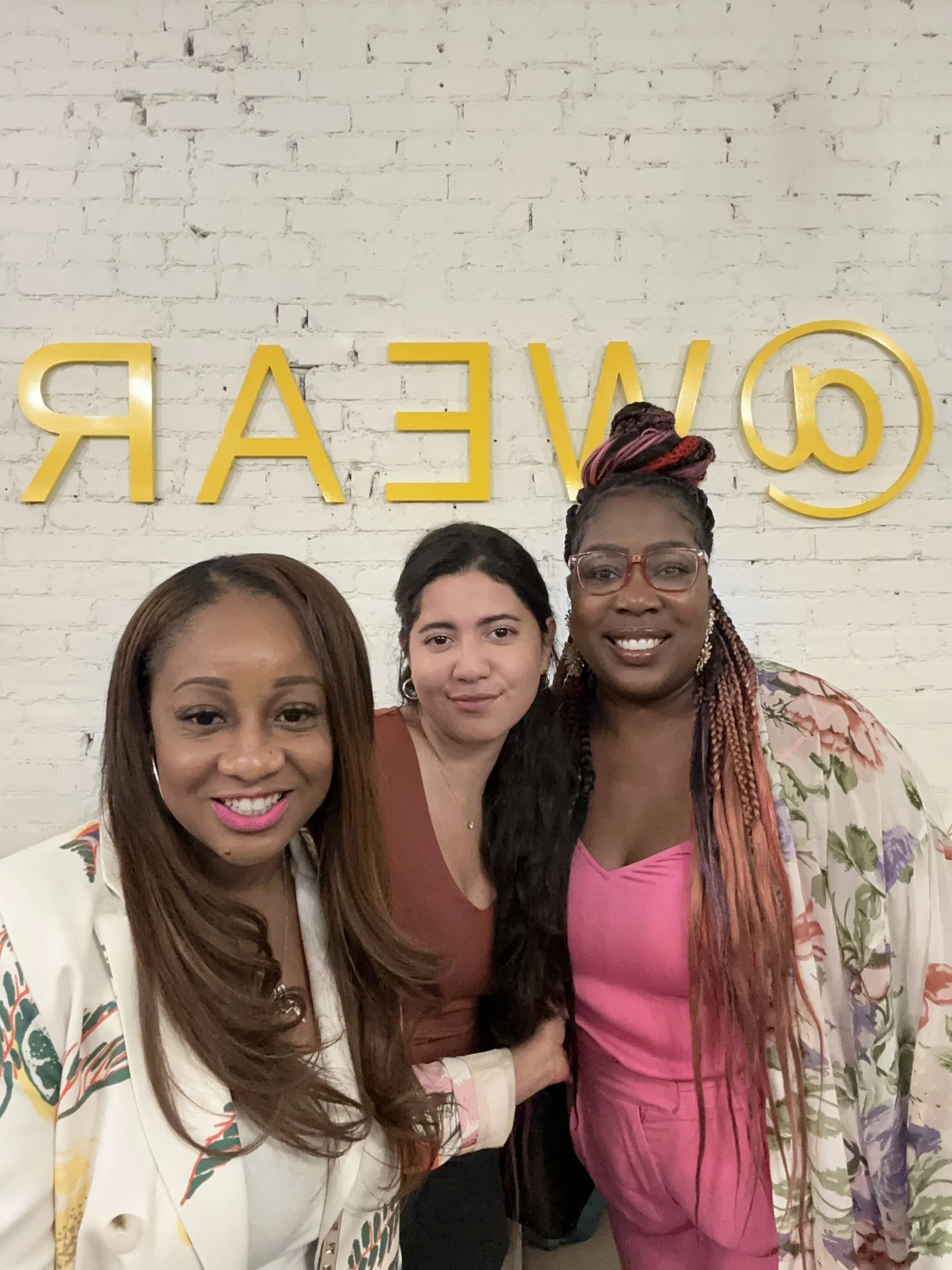 Three women smiling and posing for a photo in front of a white brick wall with yellow letters spelling 'COVEAR'.
