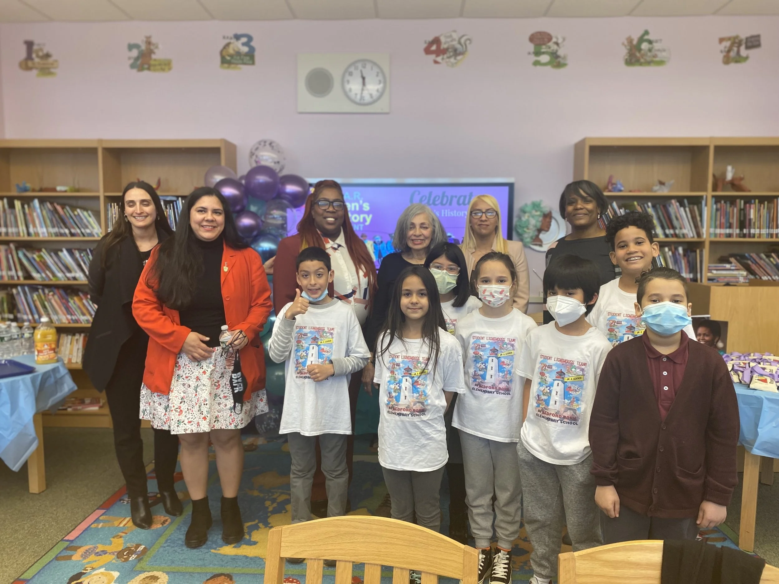 Group of children and adults smiling at a celebration event in a school library, with balloons and a colorful banner in the background.