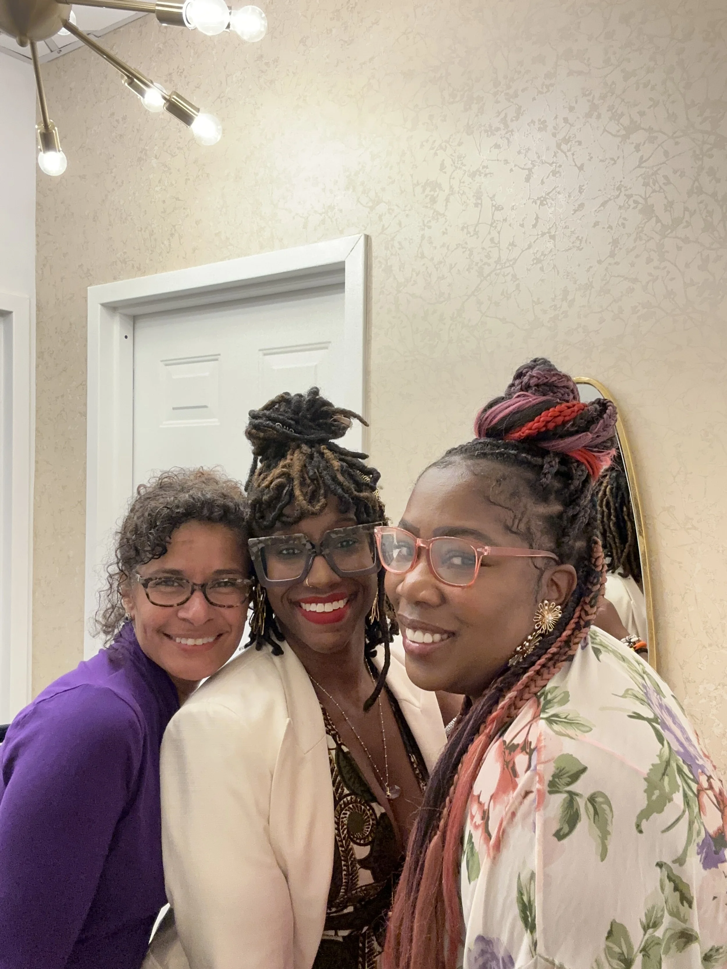 Three women smiling closely together in a room with a beige wall and a mirror. They are wearing glasses and colorful clothing, with styled hair.