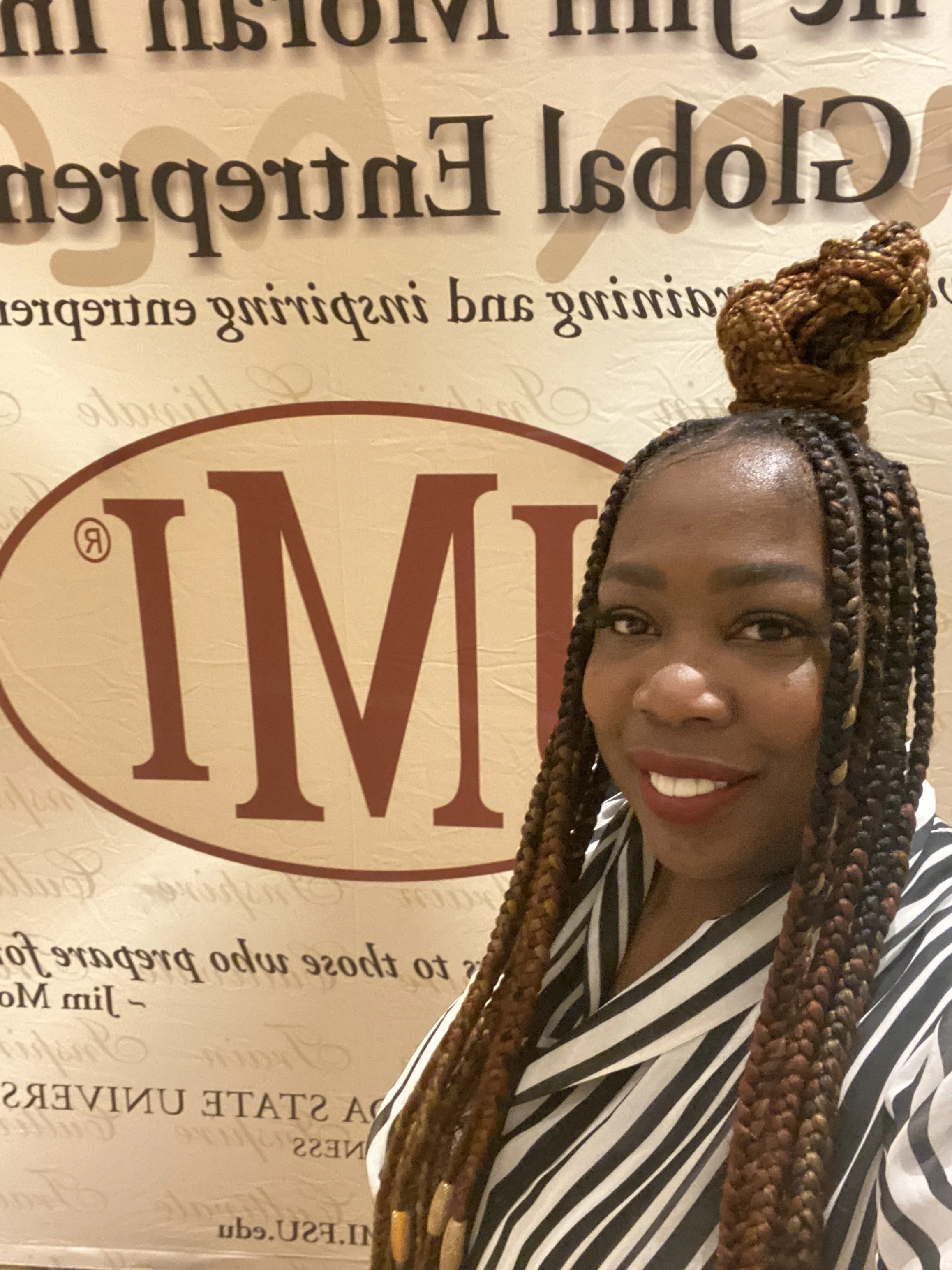 A woman with long brown braids, partly up in a bun, smiling at the camera, standing in front of a light-colored background with a large red and brown logo that reads 'MM' and some text about a steakhouse.