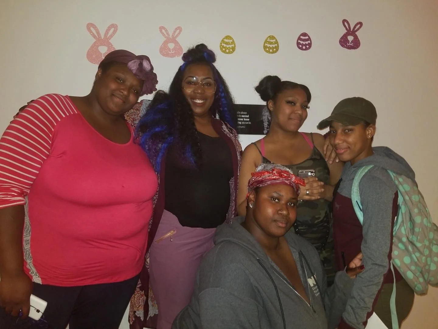 A group of six women and one girl posing together indoors, with Christmas and Easter decorations on the wall behind them.