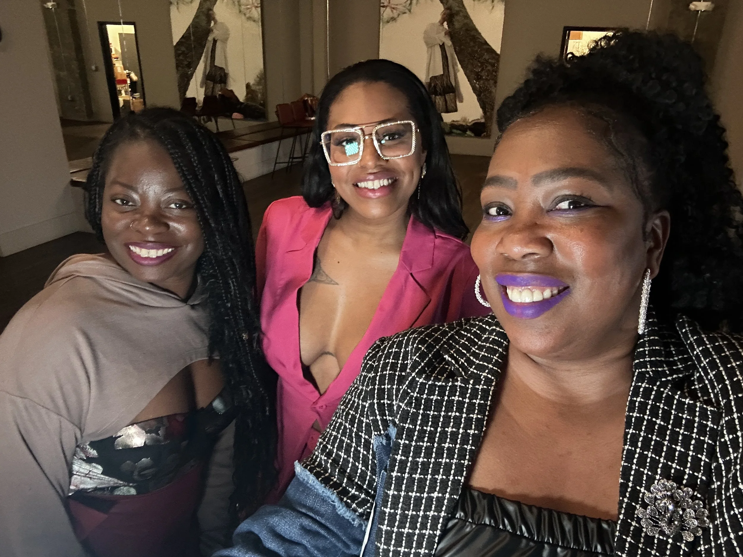 Three women smiling and taking a selfie together indoors, with colorful makeup and stylish clothing.