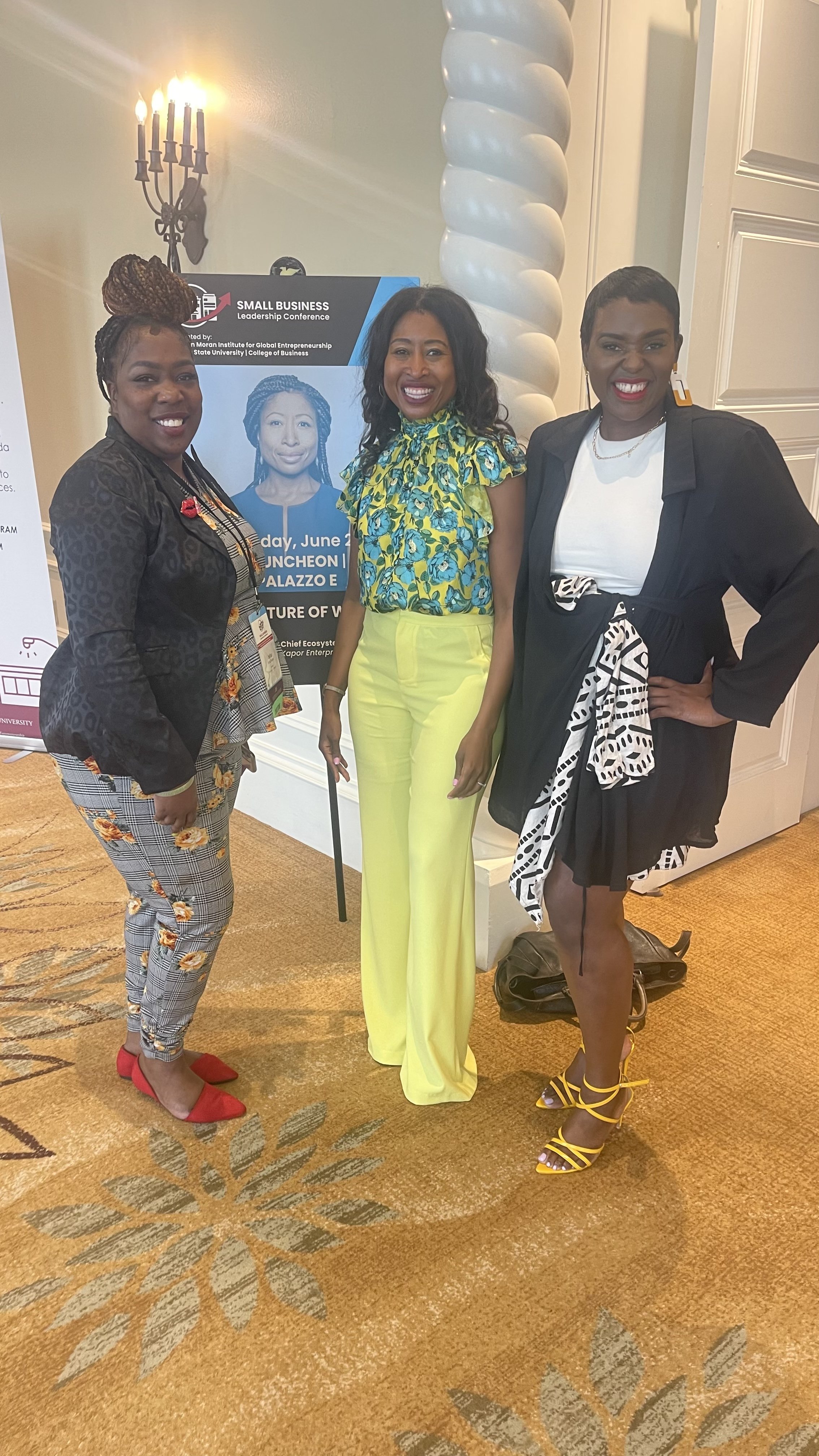 Three women standing together and smiling at an indoor conference event. One woman is wearing a black blazer with checkered pants and red shoes, the second woman is in a floral top with bright yellow pants, and the third woman is dressed in a black a