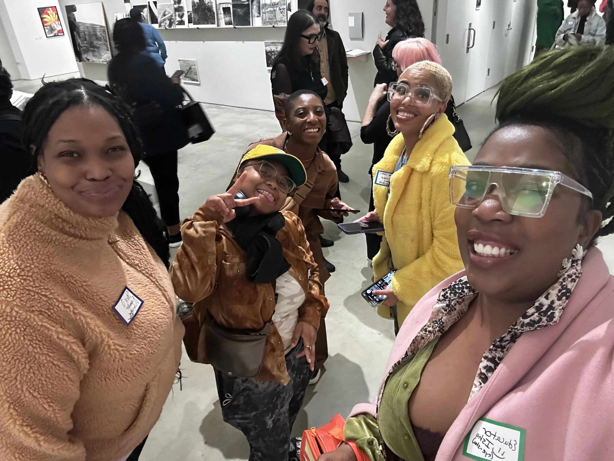 Group of five smiling women in colorful jackets at an indoor event, with art displays on the wall behind them.
