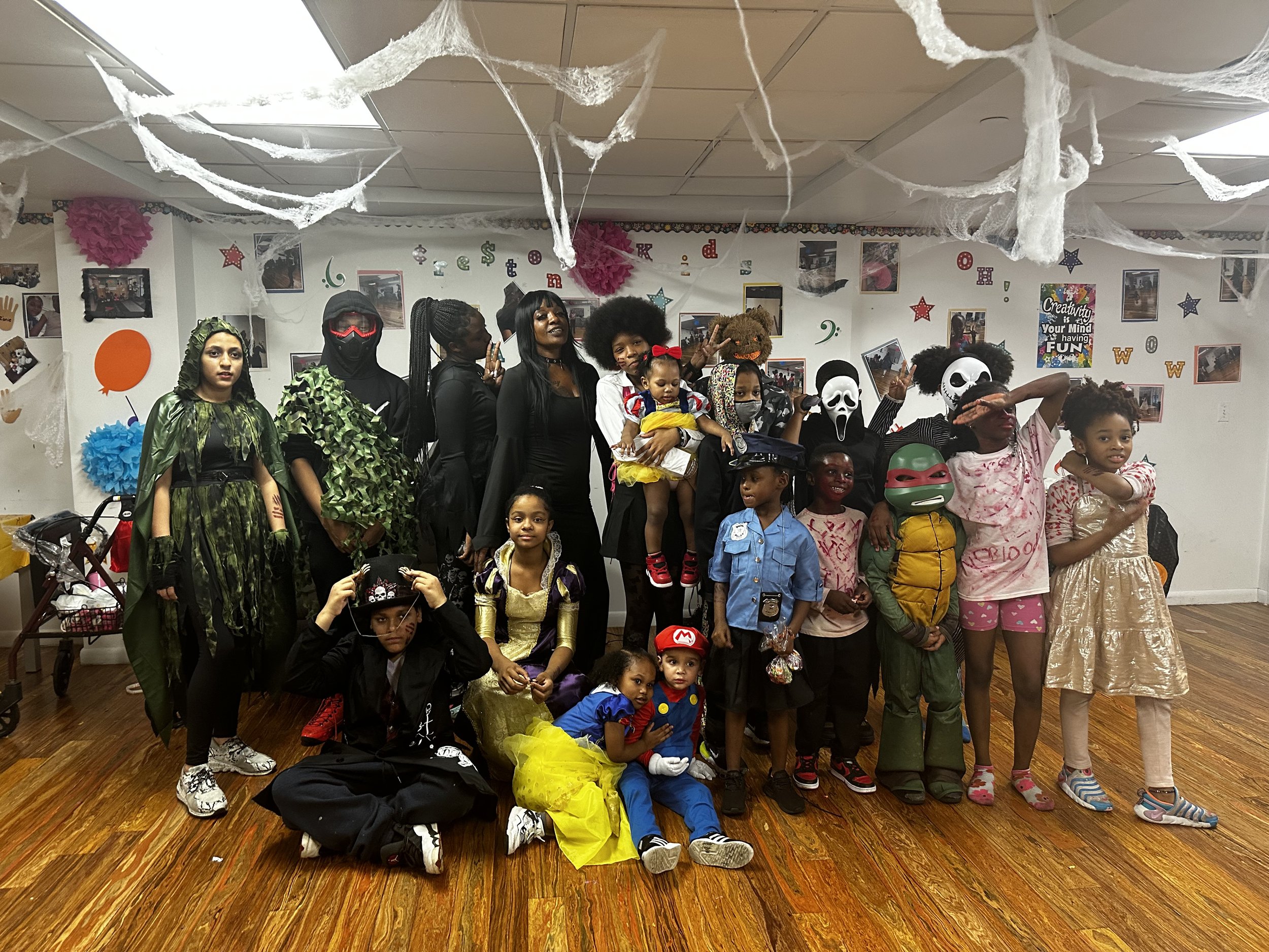 Children and adults dressed in various costumes, including superheroes, pirates, and spooky characters, posing in a decorated party room with cobweb decorations and photos on the wall.