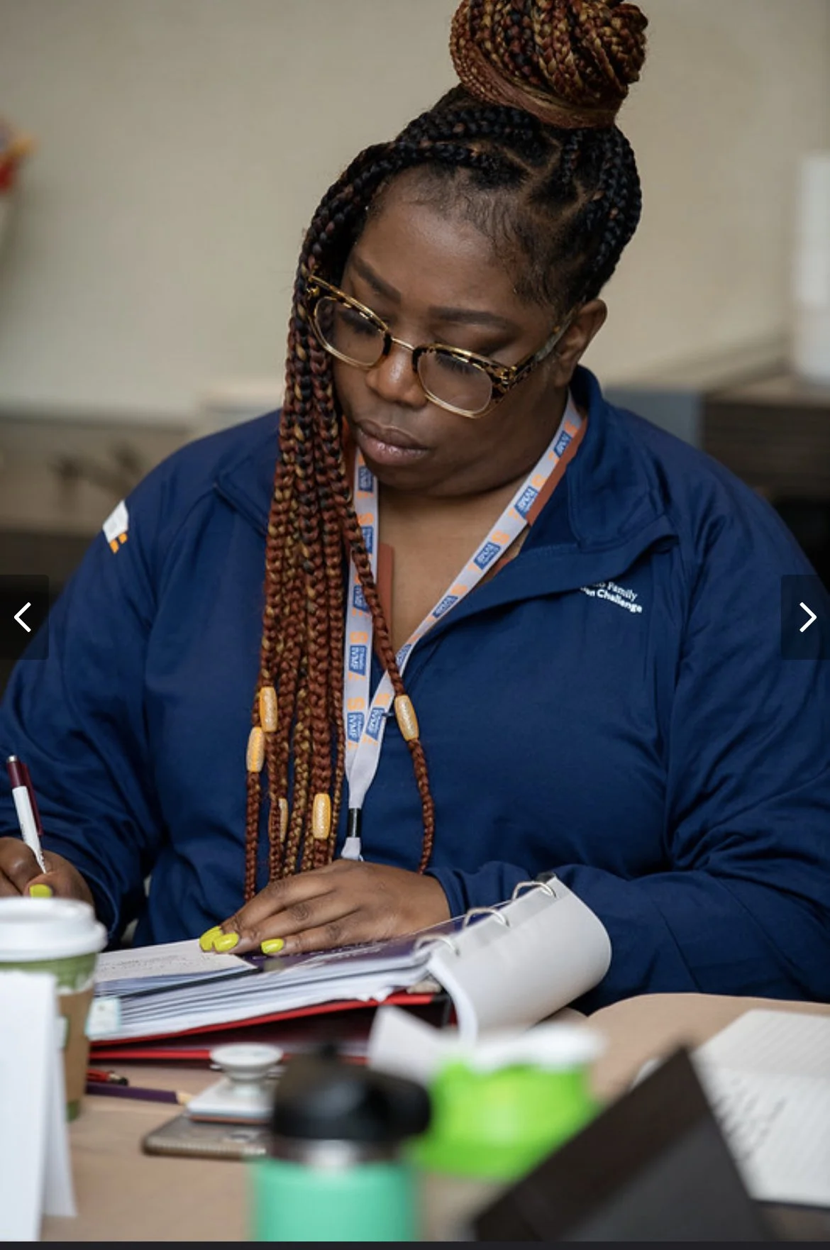 A woman with braided hair wearing glasses and a blue jacket, sitting at a table, writing in a notebook, surrounded by books, coffee cups, and other office supplies.