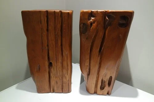 Deux sculptures en bois massives avec des surfaces lisses et des contours naturels, placées sur une surface blanche.