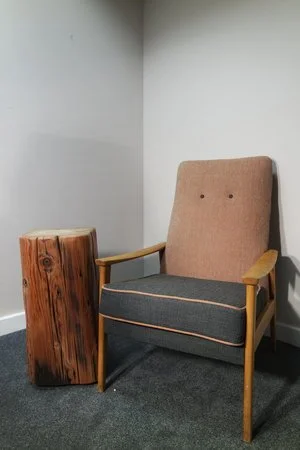 Fauteuil en tissu beige et gris avec bras en bois, à côté d'une table en bois naturel dans un coin de pièce avec murs blancs.