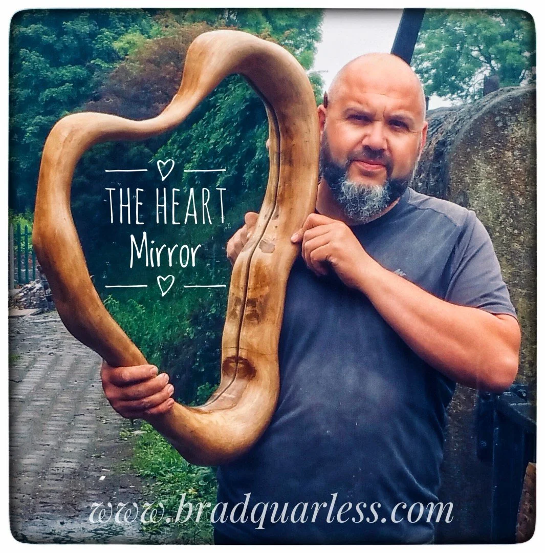Un homme avec une barbe tient un miroir en forme de cœur en bois, avec le texte "The Heart Mirror" inscrit dessus, en extérieur.