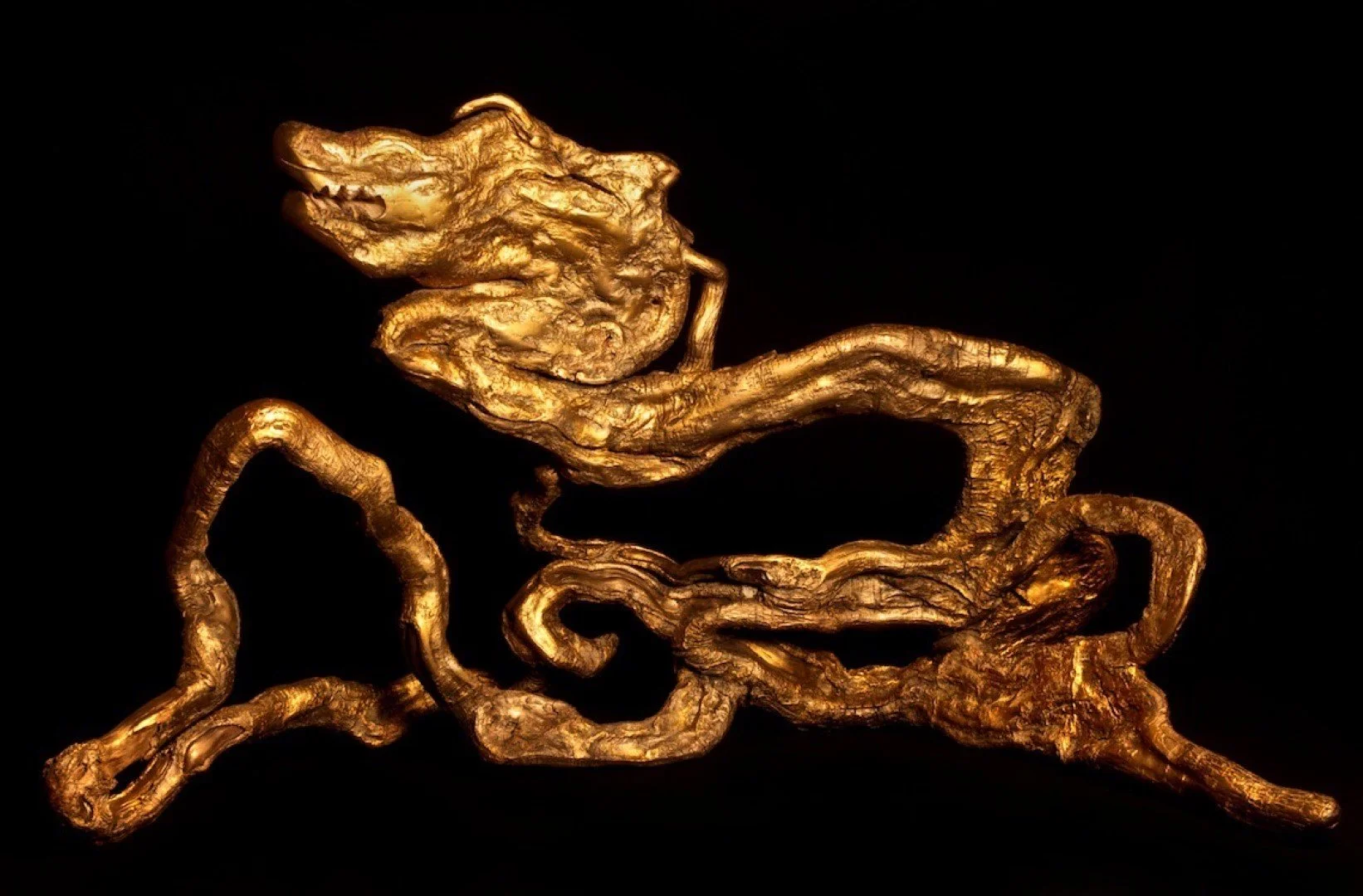 Une sculpture dorée représentant un visage humain, un animal ou une figure abstraite, avec des formes ondulantes et des textures complexes, sur un fond noir.