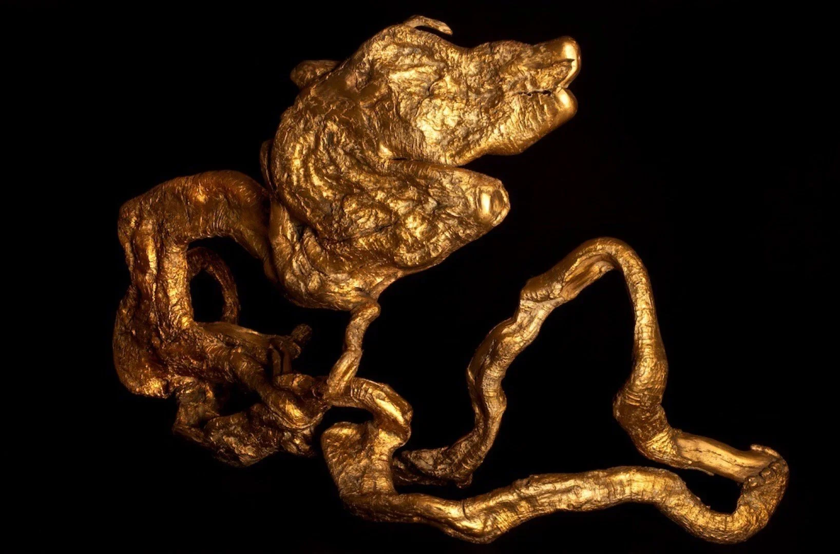 Objet artisanal en or représentant une créature ressemblant à un animal hybride ou une sculpture organique complexe, placé sur un fond noir.