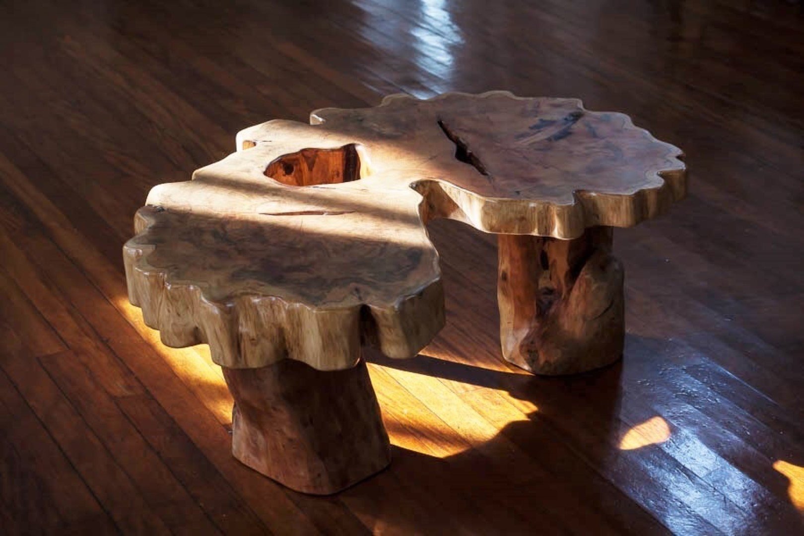 Table basse en bois brut avec une surface en forme organique et deux piètements en bois massif, posée sur un sol en bois.
