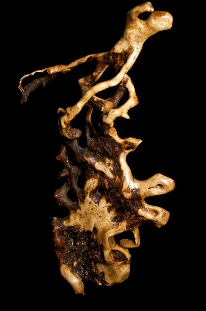 Une sculpture abstraite en bois sculpté, représentant un visage humain stylisé, avec des contours fluides et organiques, sur fond noir.