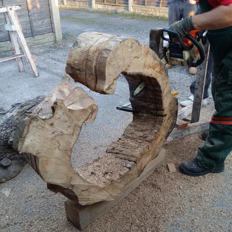 Un bûcheron sculpte une pièce de bois massive avec une tronçonneuse, entouré de copeaux de bois.