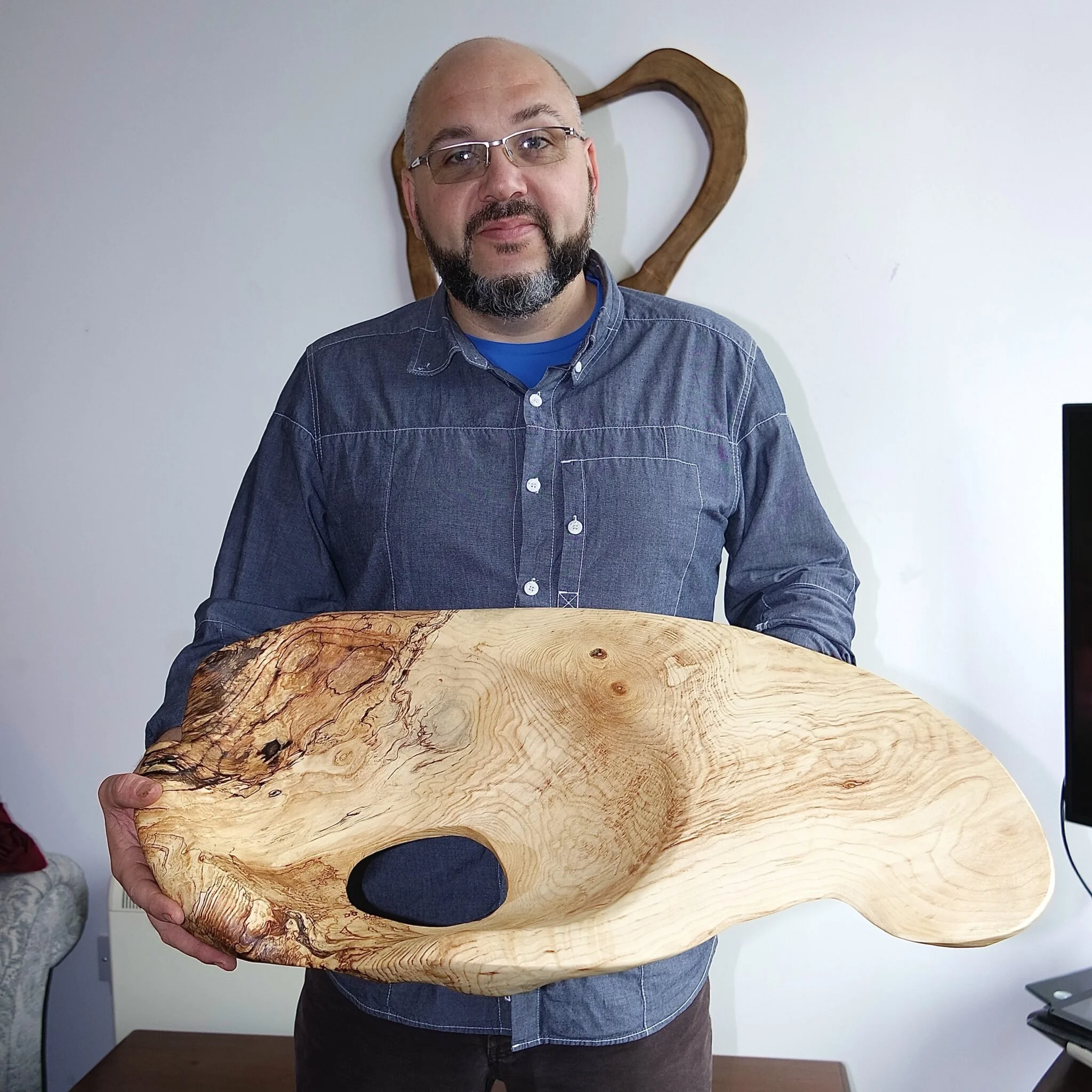 Un homme tient une sculpture en bois sculpté de forme organique, avec des motifs naturels et des textures variées, dans une pièce avec un mur blanc et un canapé visible.