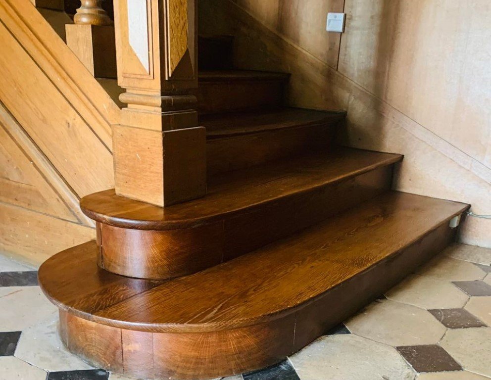 Escalier en bois poli avec une rampe en bois, situé à côté d'un mur en pierre de couleur beige.