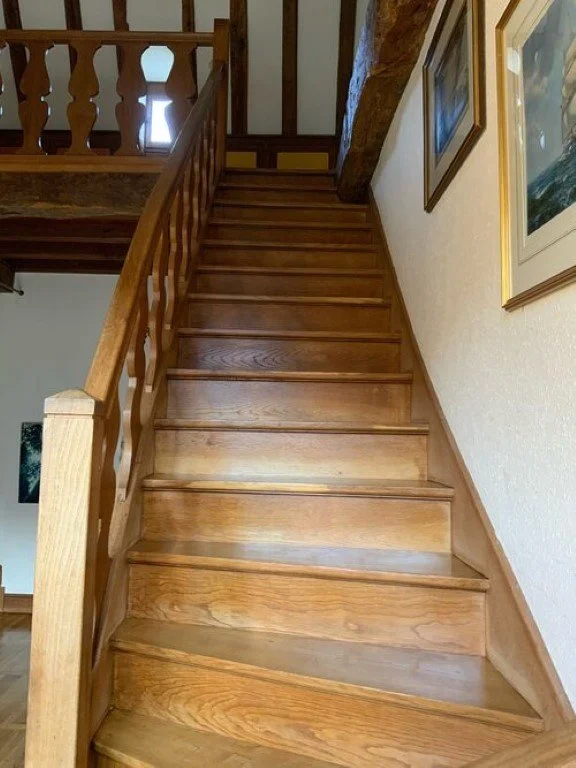 Escalier en bois menant à un étage supérieur dans une maison, avec des tableaux accrochés au mur à droite.