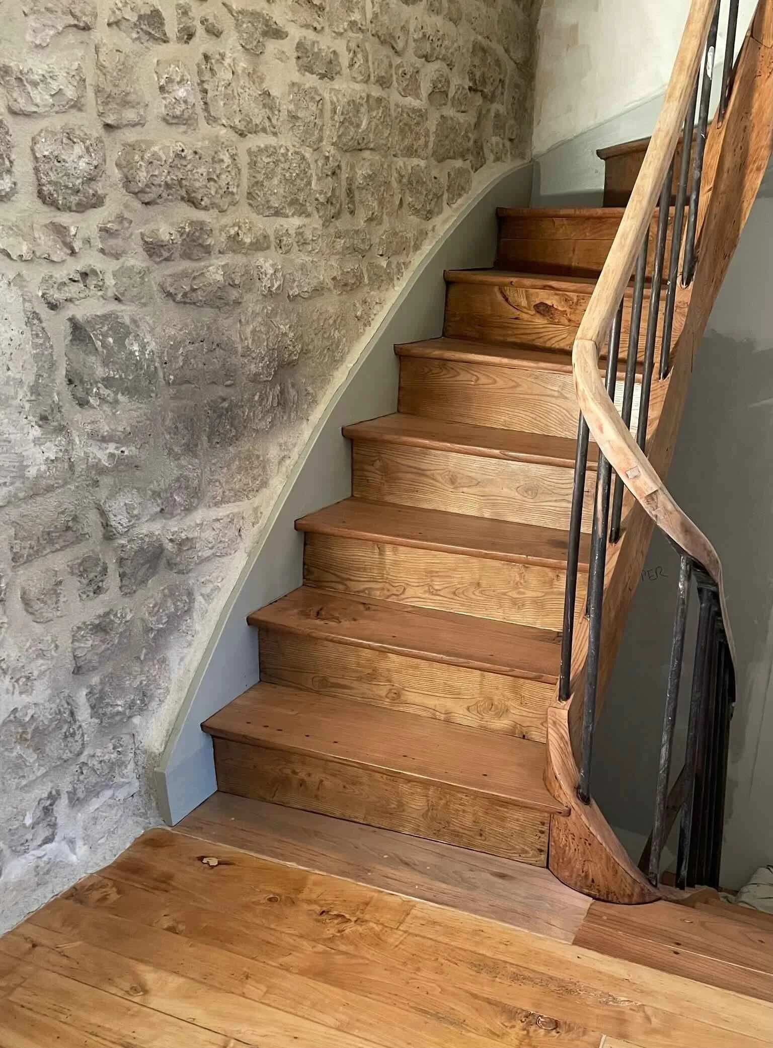 Escalier en bois avec main courante en métal, murs en pierre et sol en bois massif.