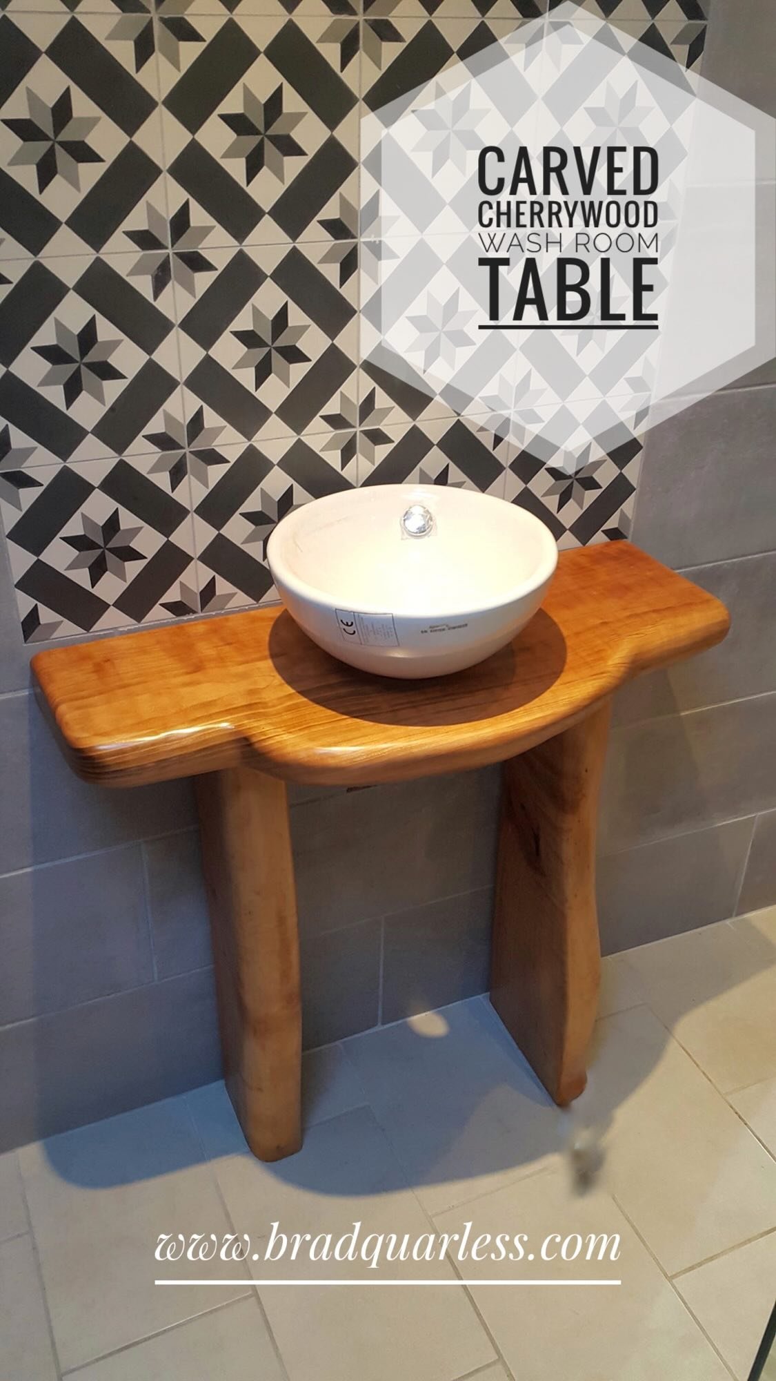 Un lavabo en céramique blanc sur une table en bois, en arrière-plan un mur avec des carreaux de style géométrique noir, blanc et gris, avec un panneau indiquant un nom de chambre et une table.
