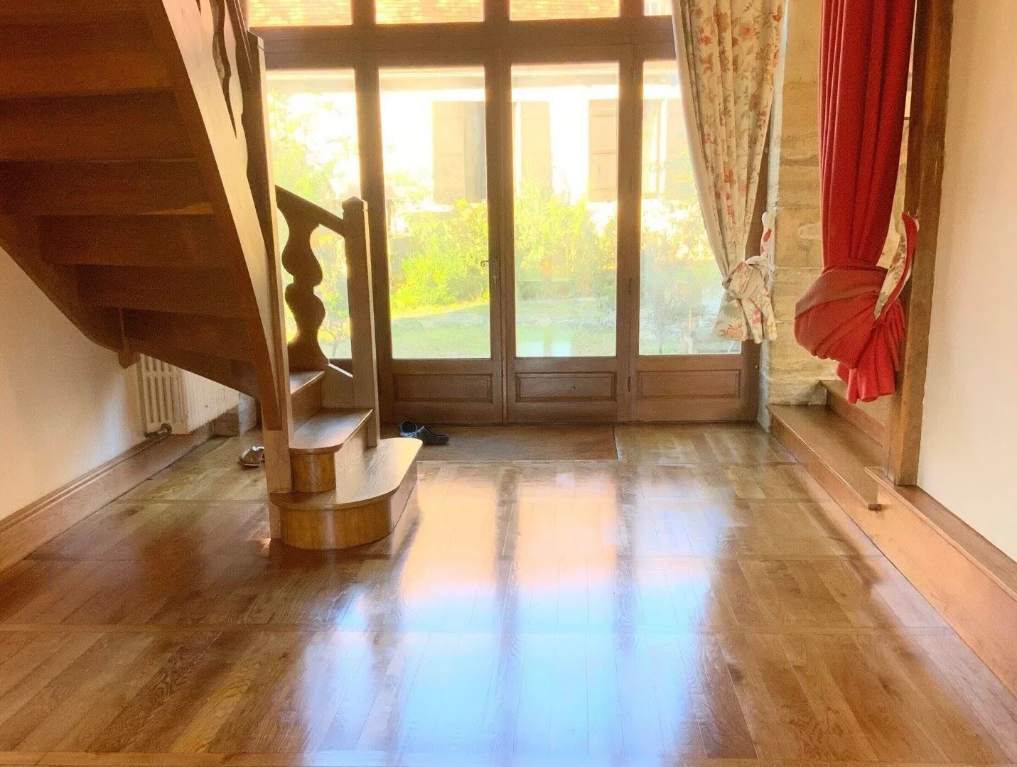 Intérieur d'une pièce avec un sol en bois, un escalier en bois à gauche, des portes-fenêtres en verre avec rideaux, et un mur en pierre derrière.