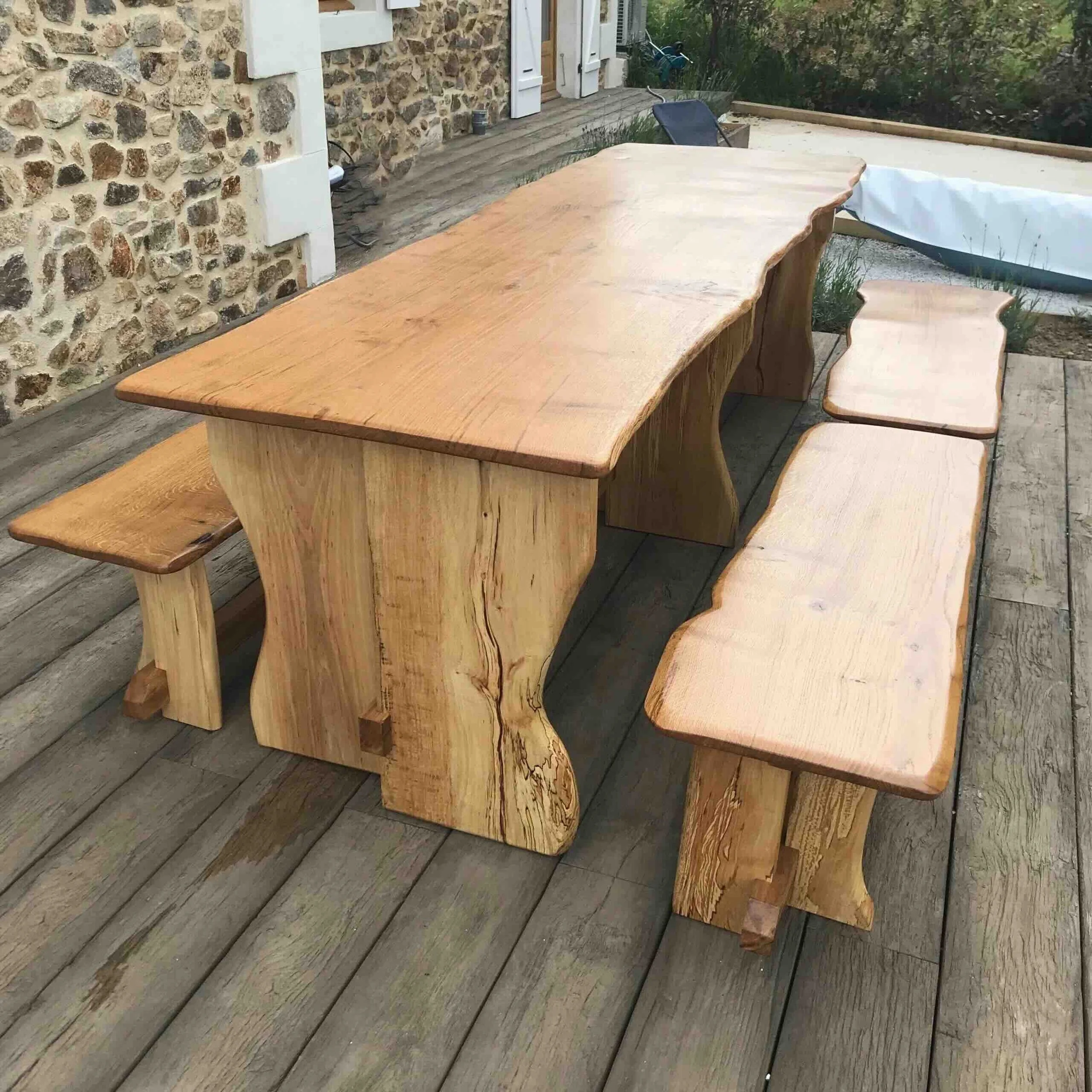 Table en bois naturel dans un espace extérieur avec un mur en pierre et du mobilier de jardin en arrière-plan.