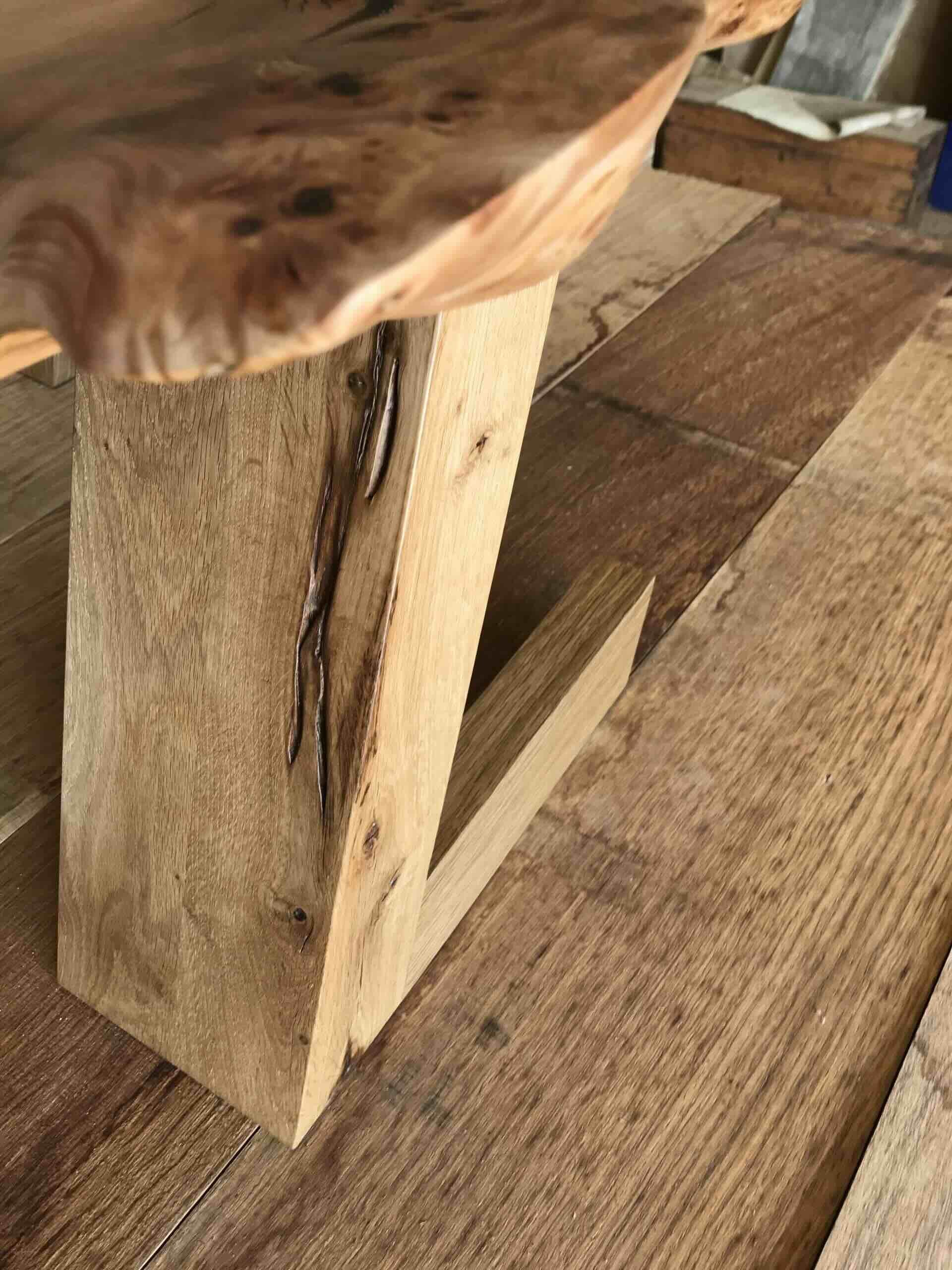 Vue d'une table en bois brut avec un espace creux en dessous et des planches de bois posées à côté, sur un sol en bois.
