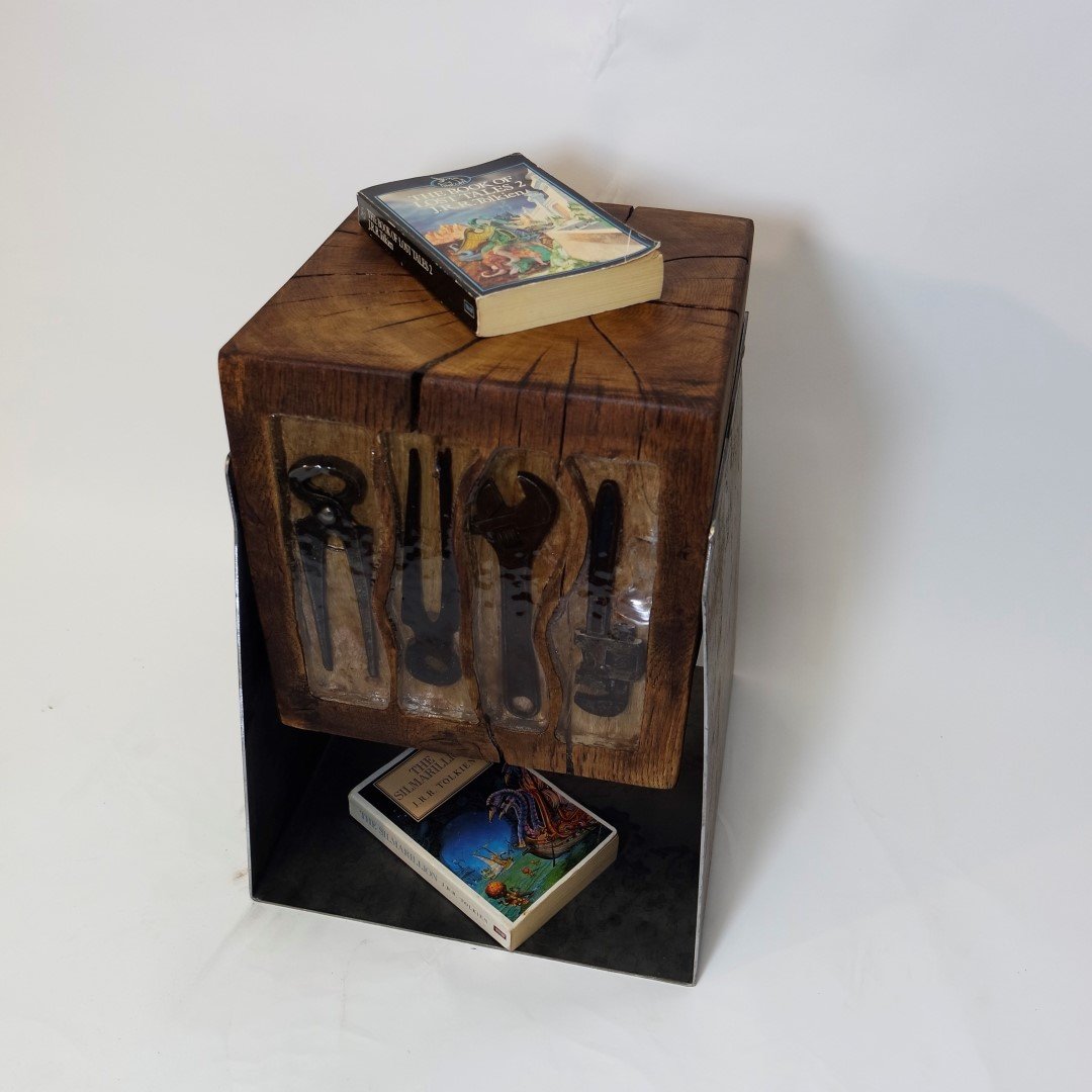 Une petite table en bois avec un livre dessus et un autre livre au bas de la table. La table a une surface en bois brut avec des fissures et des ustensiles en métal derrière un panneau en plastique fixé au bois.