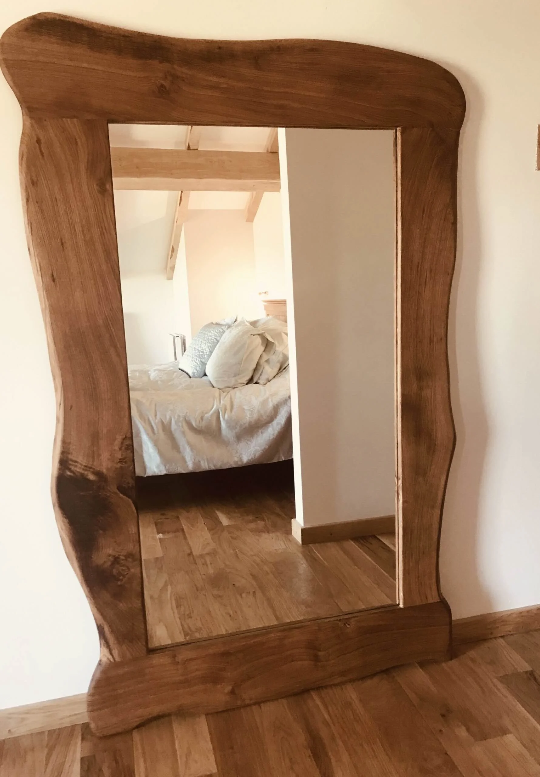 Miroir en bois avec cadre courbé, reflétant un lit avec des oreillers blanc et une pièce en bois.