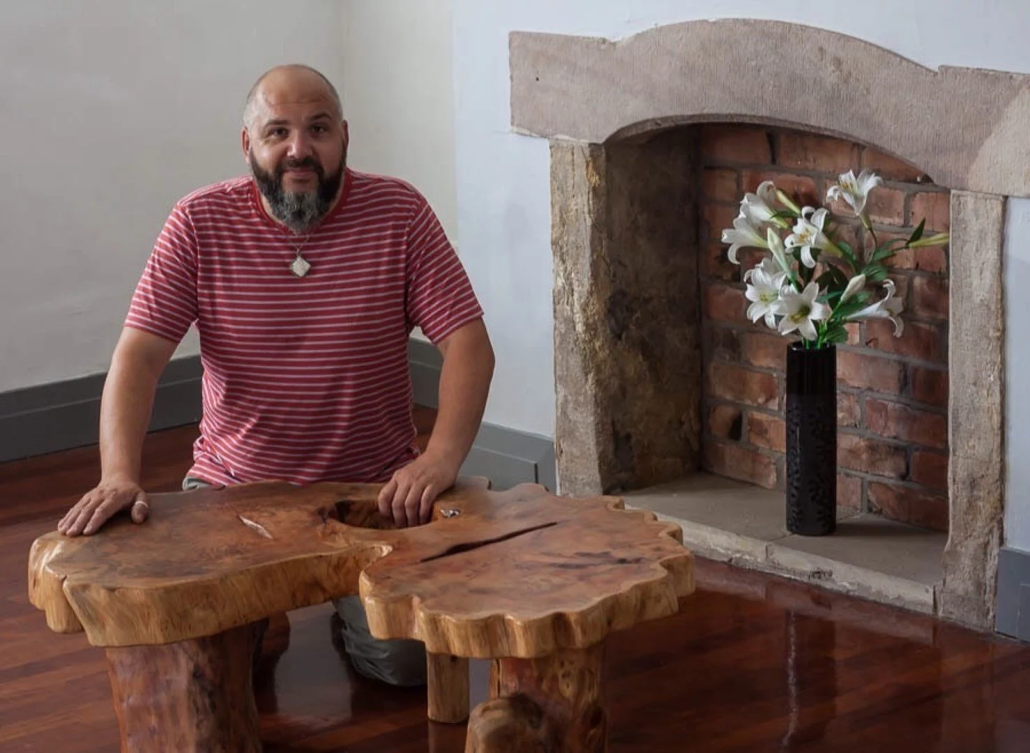 Un homme avec une barbe et un t-shirt rayé rouge et blanc est assis à une table en bois massif, à côté d'une cheminée en brique avec une vase noire contenant des fleurs blanches.