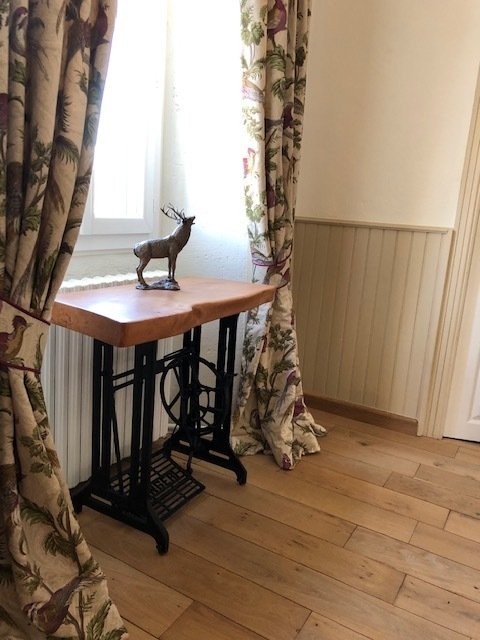 Une table en bois avec une machine à coudre ancienne en dessous, près d'une fenêtre avec des rideaux à motifs floraux, et une petite statue de renne sur la table.