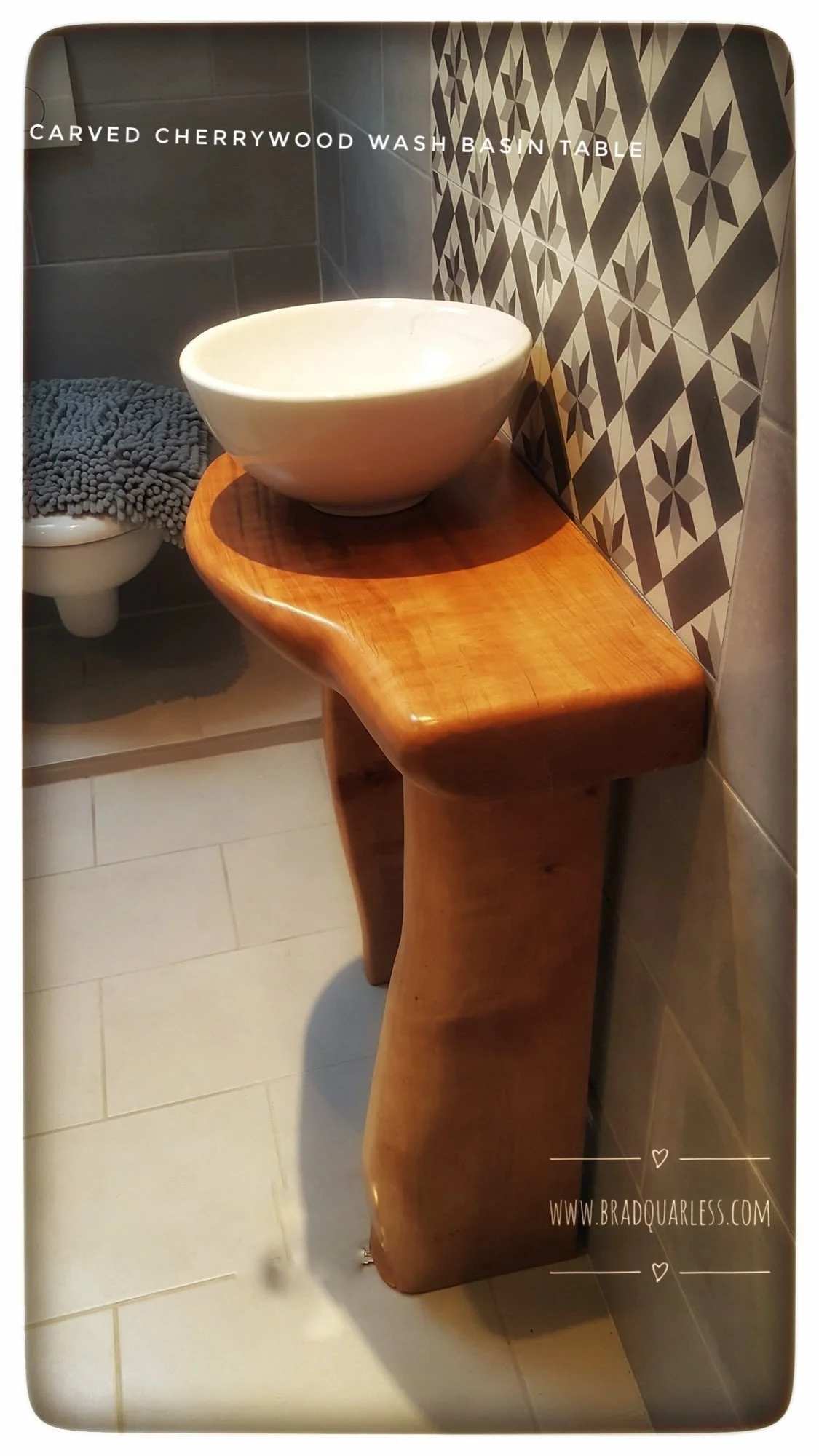 Un lavabo en céramique blanc rond posé sur une table en bois pivotante, dans une salle de bain avec des murs en tuiles décoratives et un toilette avec un tapis de bain gris à côté.