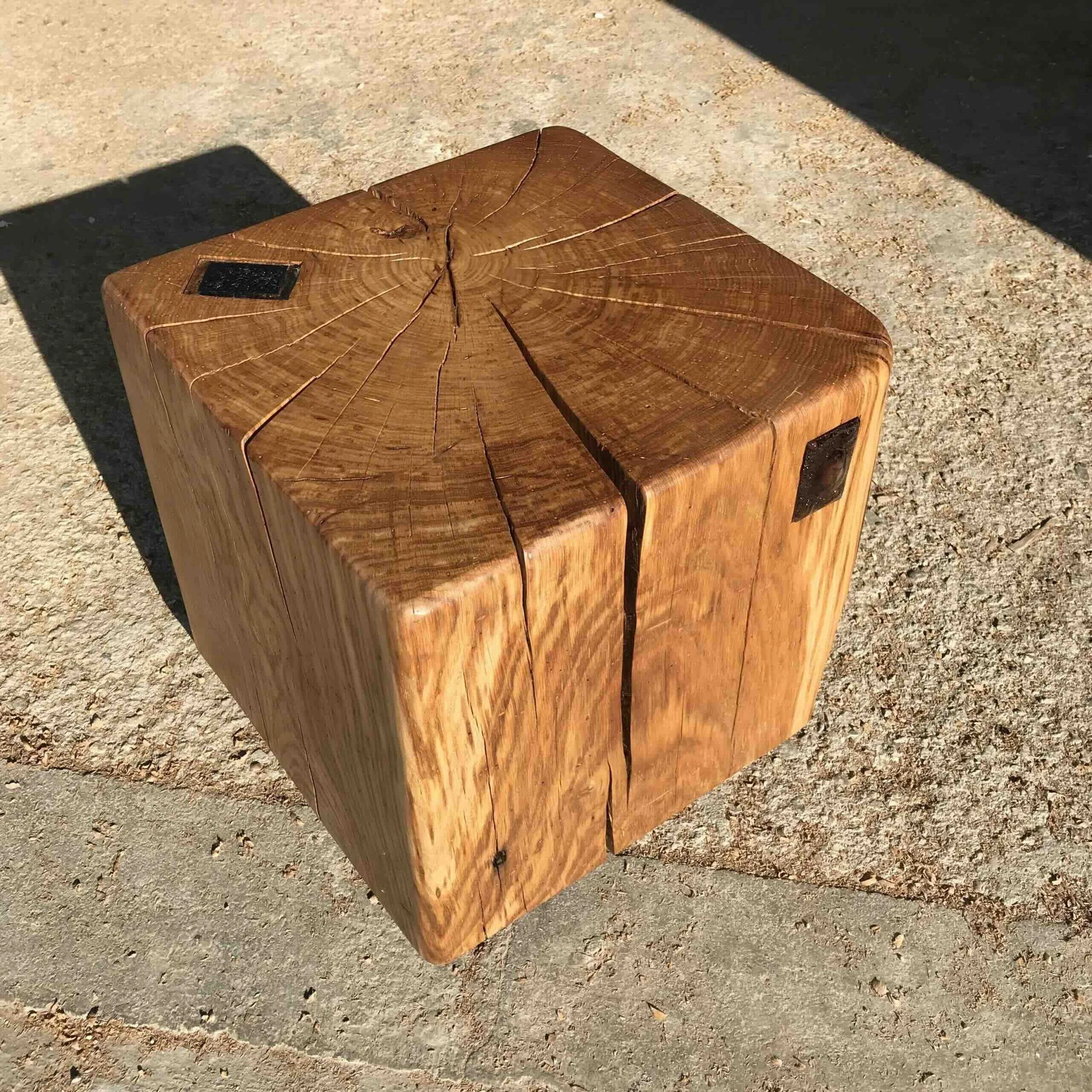 Une table en bois naturel avec une surface lisse, présentant des fissures visibles et une plaque noire fixée en haut à gauche. La surface est éclairée par la lumière naturelle, créant des ombres sur le sol en béton.