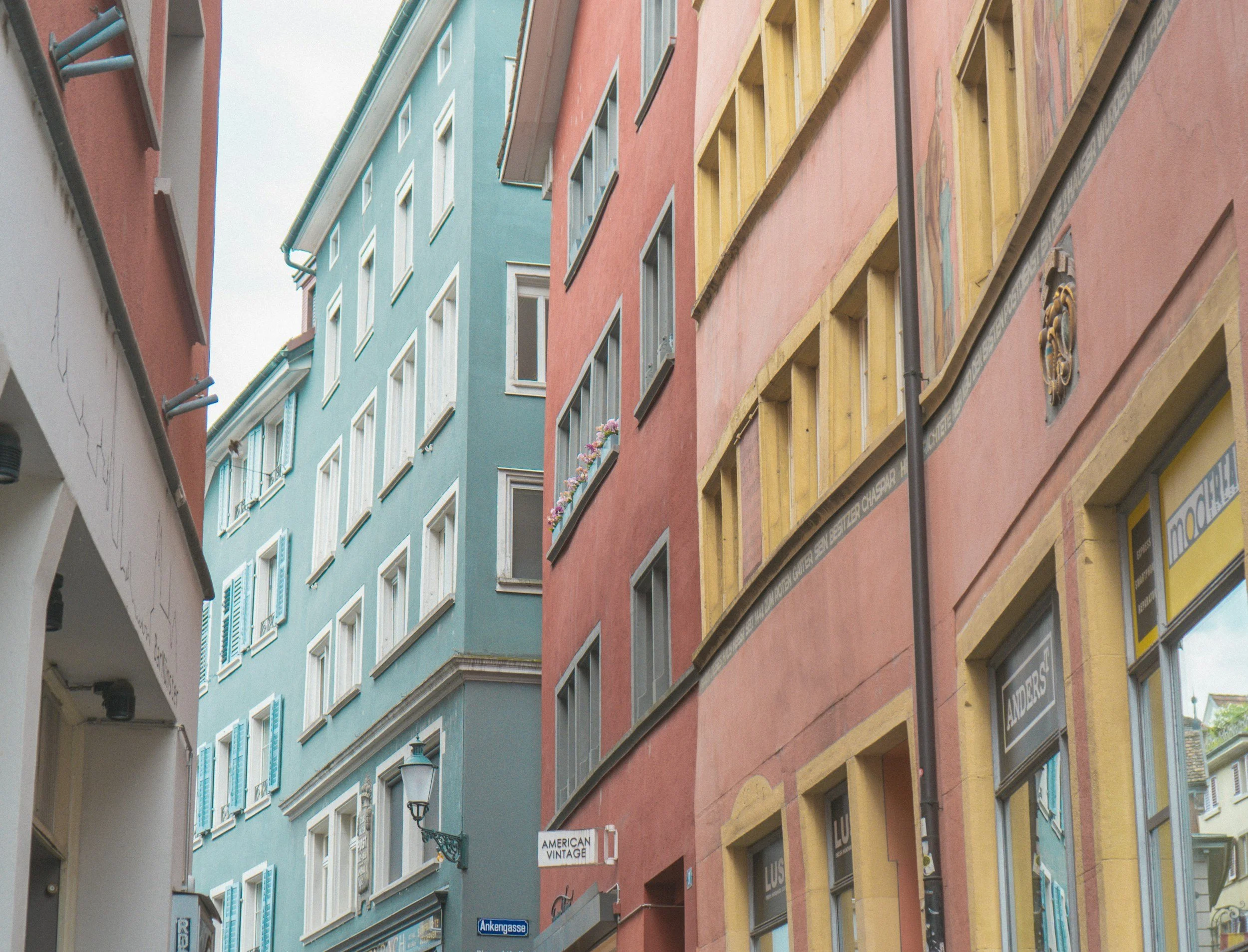 Vista di stretta strada urbana con edifici colorati in toni pastello, alcuni con vasi di fiori alle finestre, e cartelli di negozi.