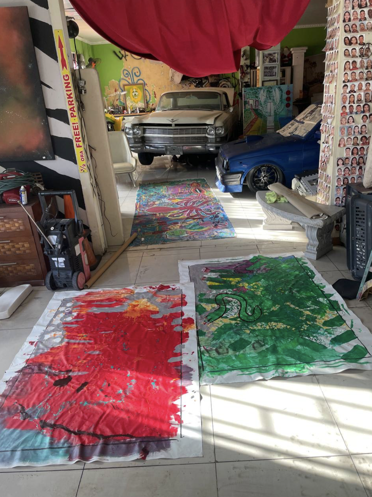 Photo of an indoor space with two large, colorful abstract paintings on the floor, a vintage white car and a blue toy car, mural art, numerous small photos on the right wall, and various furniture and art supplies.