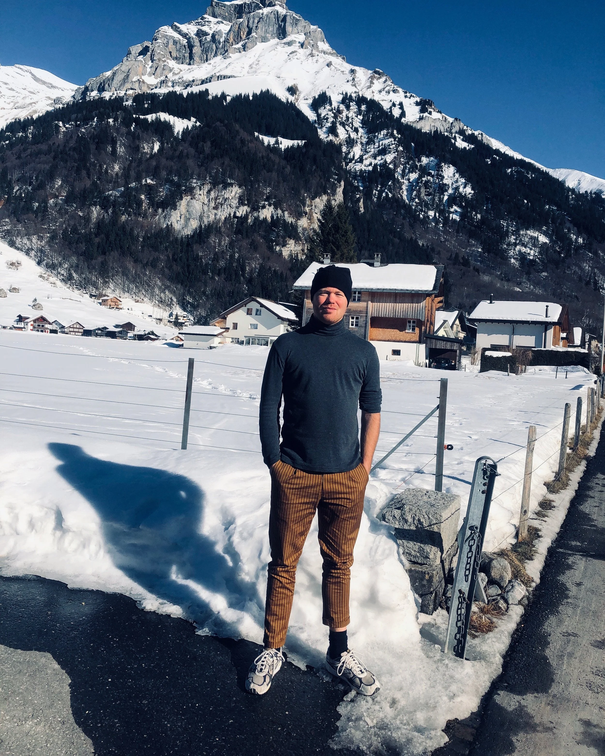 A man standing on a snowy mountain landscape with snow-covered houses in the background. He is wearing a black beanie, a dark long-sleeve shirt, brown striped pants, and sneakers. There is a large mountain with snow and trees behind him, and part of a ski or snowboard is visible on the snow.