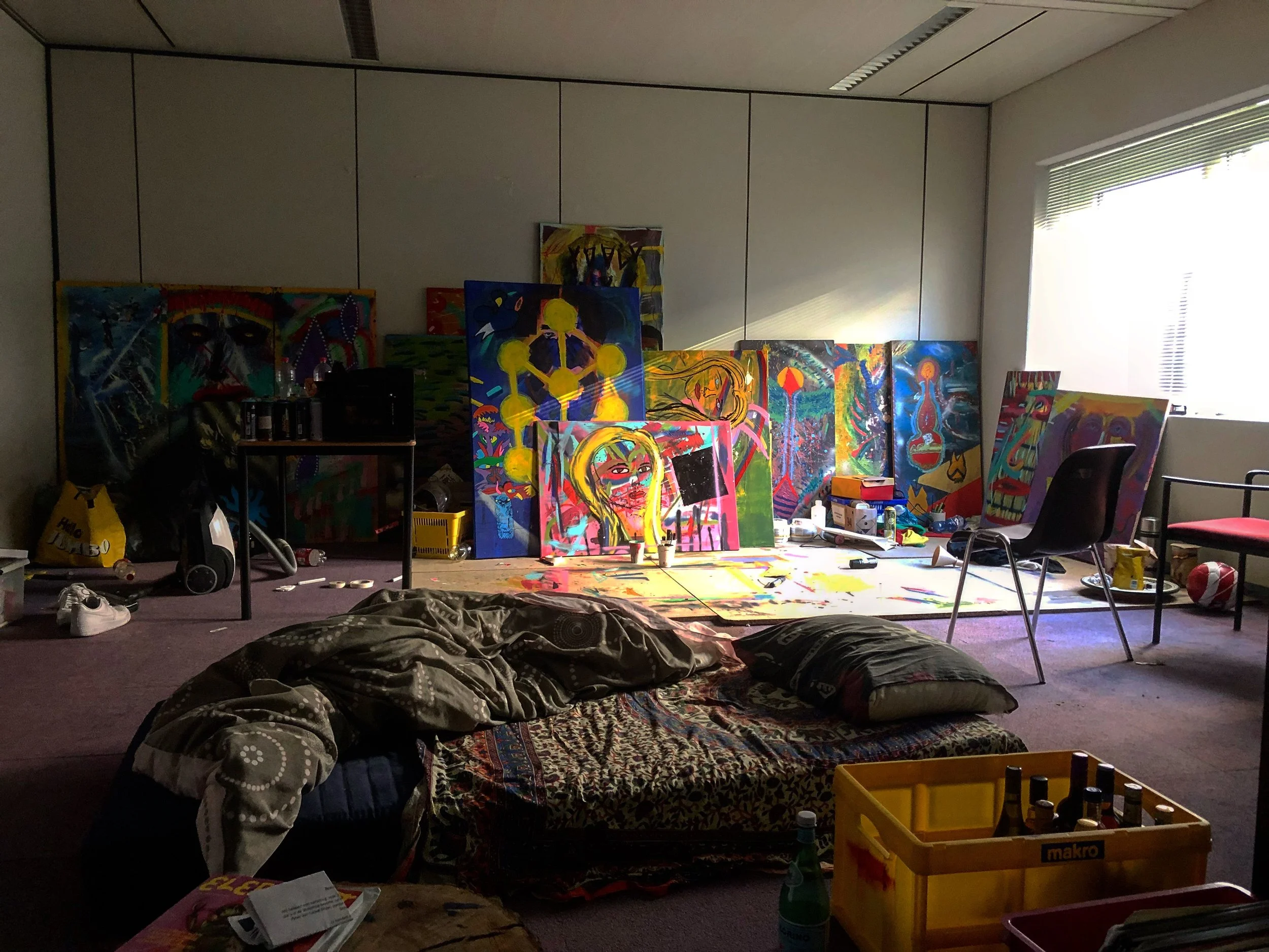 An art studio with numerous colorful paintings on the wall and floor, art supplies scattered around, and a window letting in natural light.